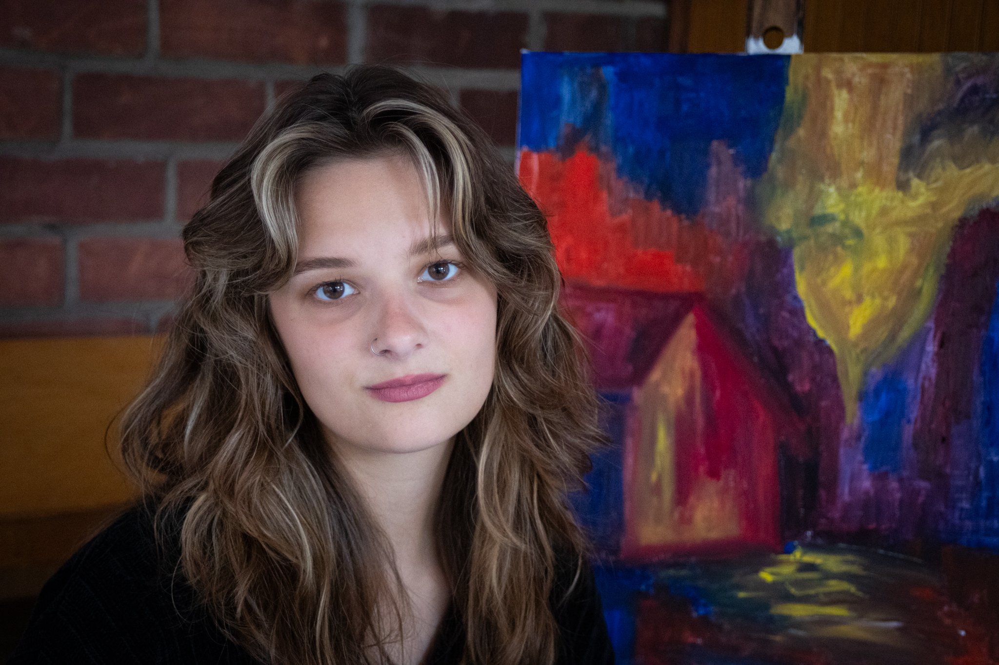 A young woman with brown, wavy hair and a nose piercing standing in front of a colorful abstract painting.