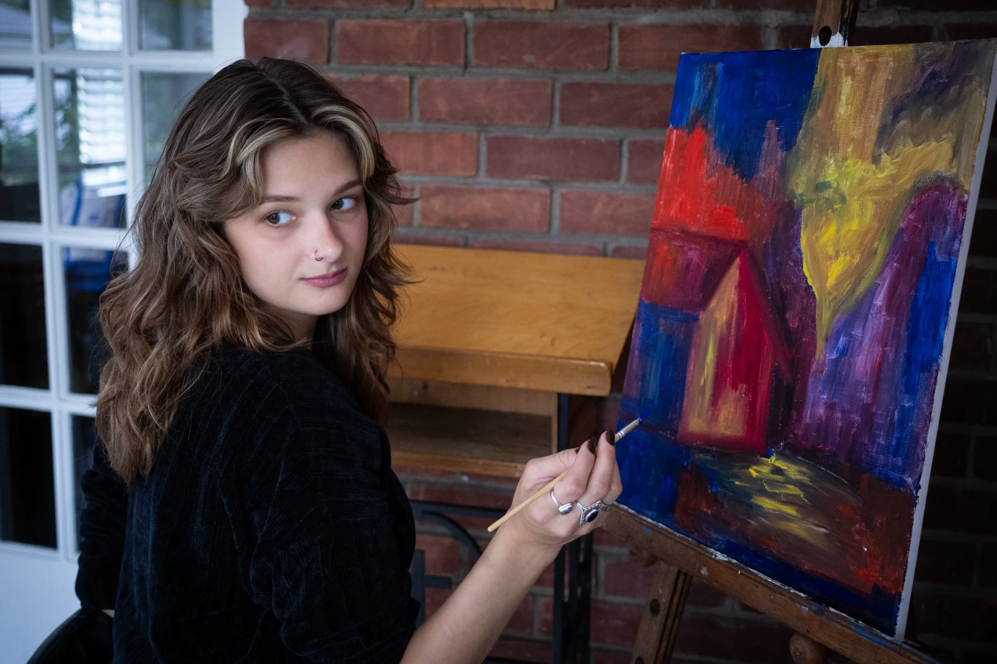 A young woman with wavy hair and a nose piercing painting an abstract art piece on canvas with bright colors including red, yellow, blue, and purple, while sitting in front of a brick wall.