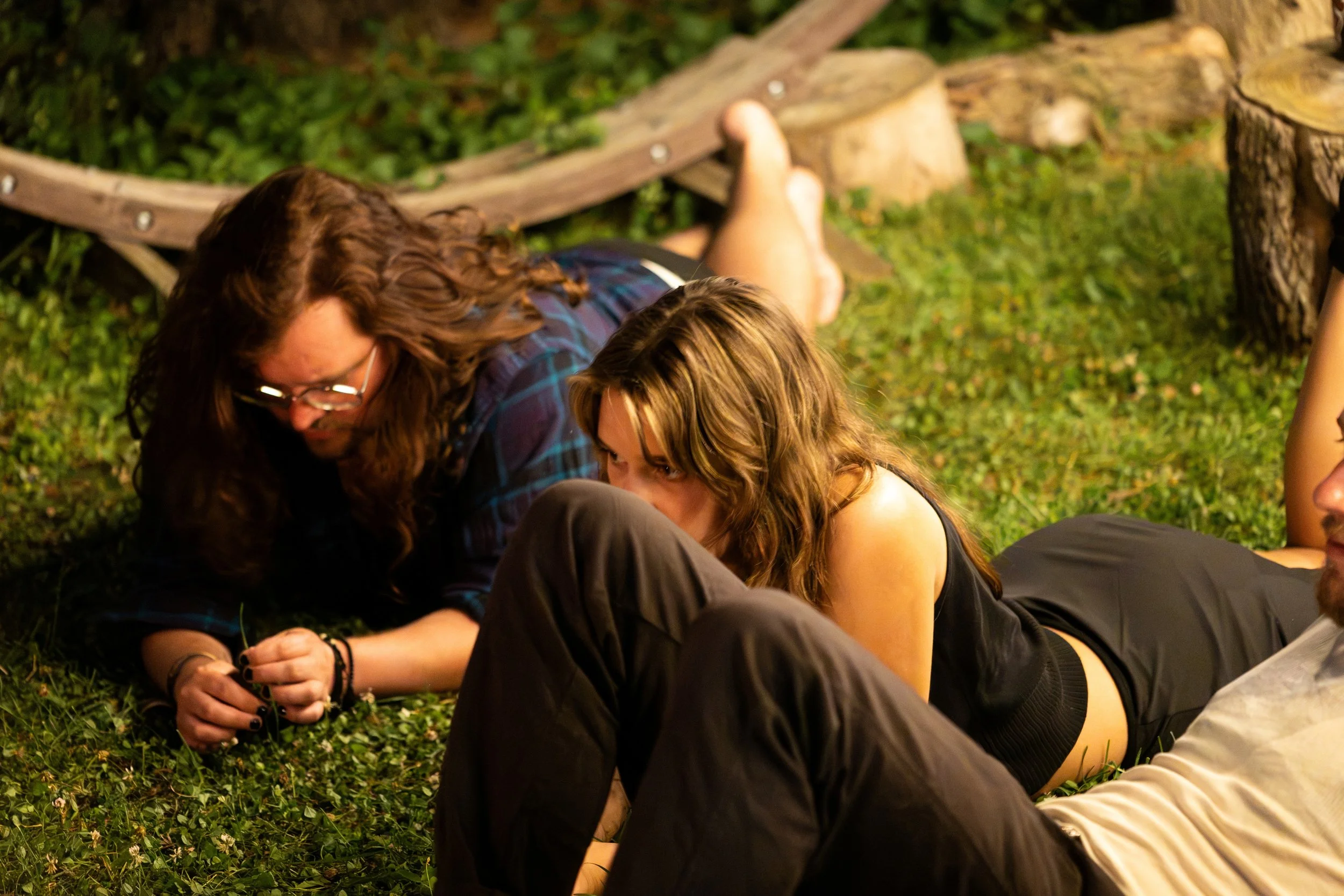 Two people with long hair, one with glasses, sitting on grass next to a wooden log with greenery behind them. Both appear to be engaged in an activity together outdoors.