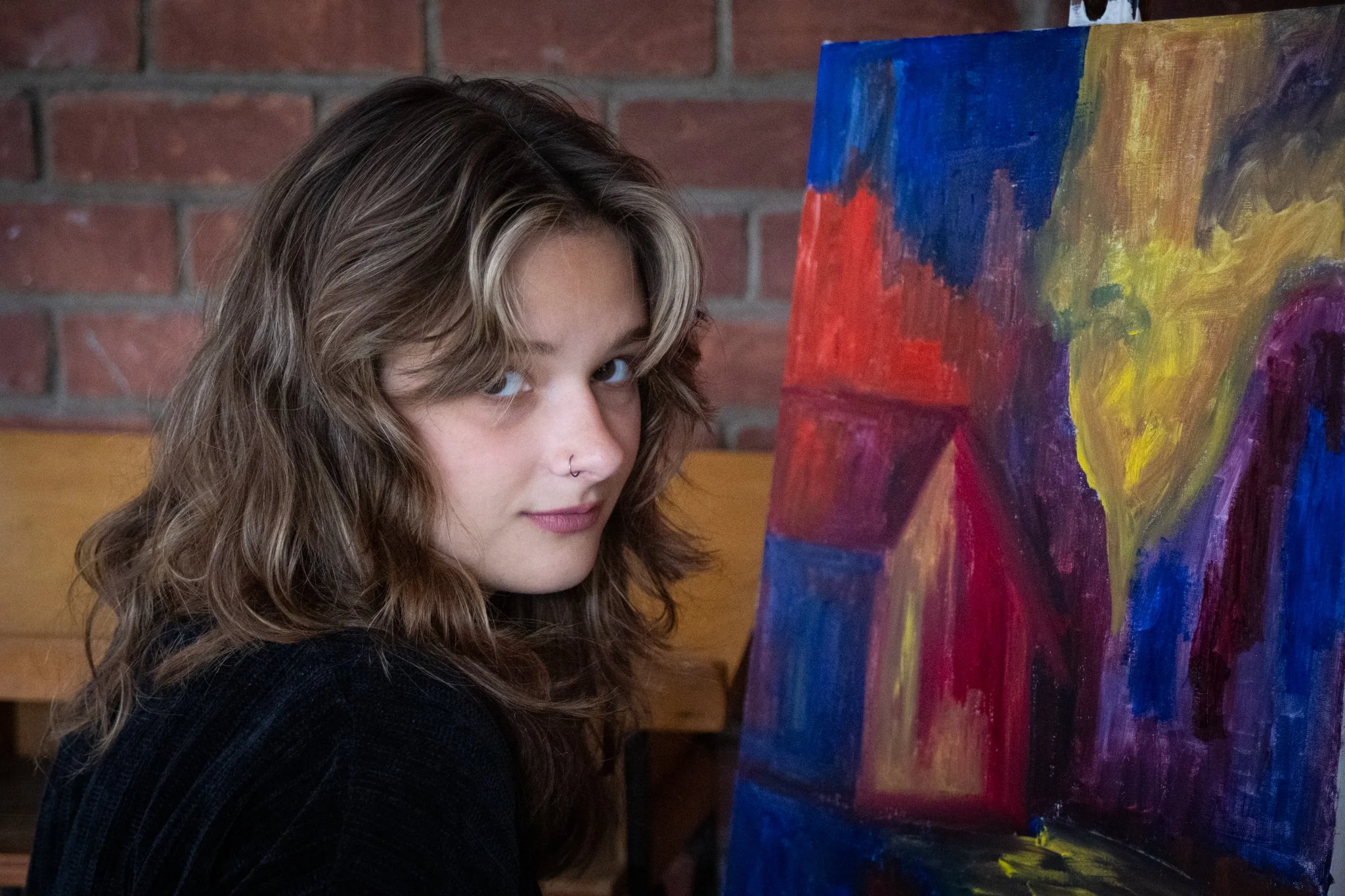 A young woman with wavy, shoulder-length hair and a nose ring looking at a colorful abstract painting on a canvas, standing in front of a brick wall.