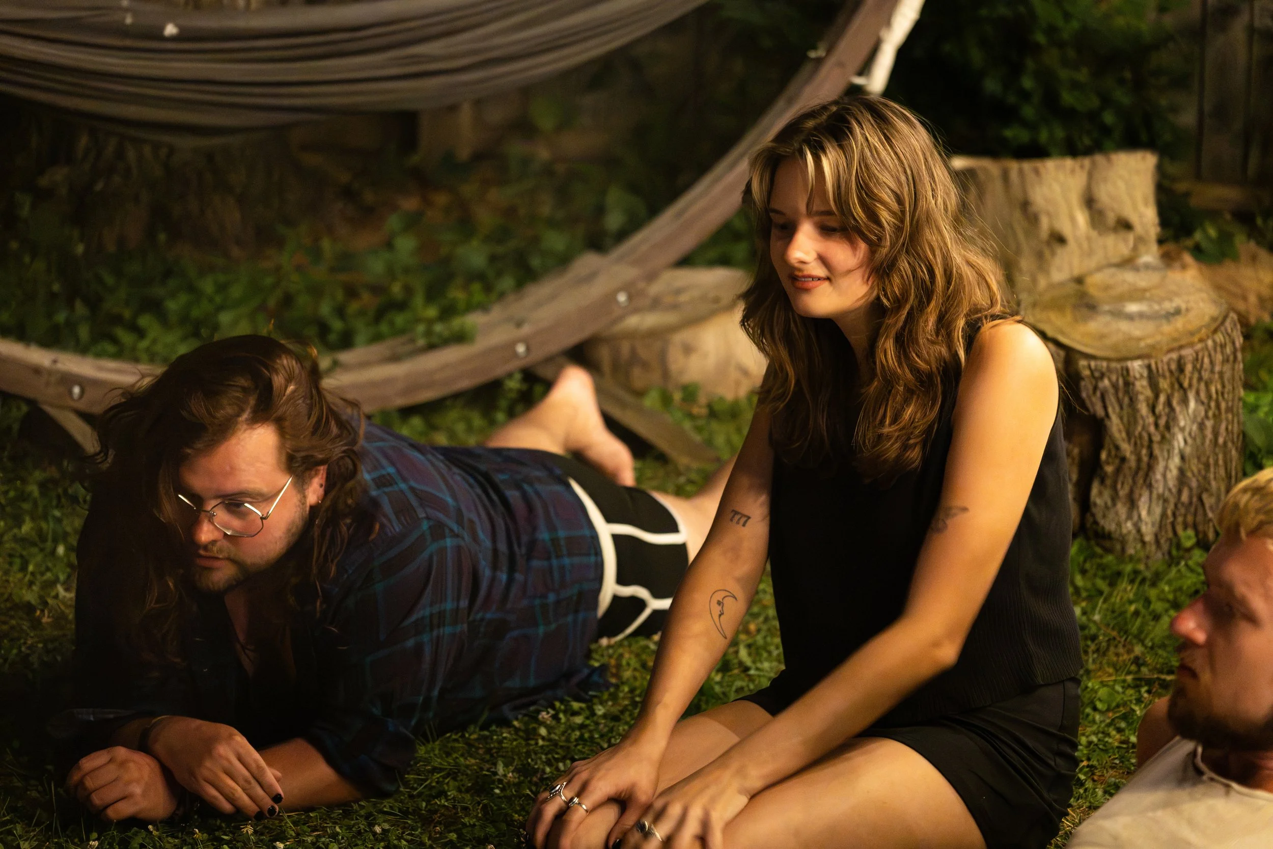 A woman with wavy, shoulder-length hair and tattoos on her arm is sitting outside at night, smiling and looking down. She is wearing a black sleeveless top. Next to her, a man with long hair, glasses, and a beard is lying on the ground, also looking down. The background features greenery, a tree stump, and a wooden structure.