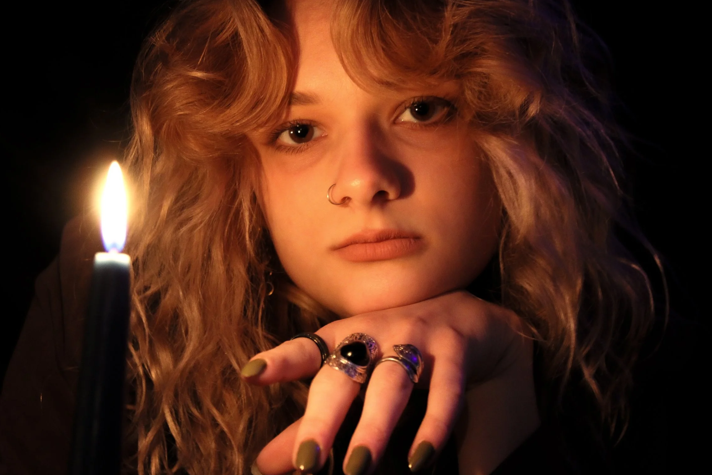 A woman with long wavy hair and multiple rings on her fingers gazes into the camera, with a lit candle in the foreground illuminating her face against a dark background.
