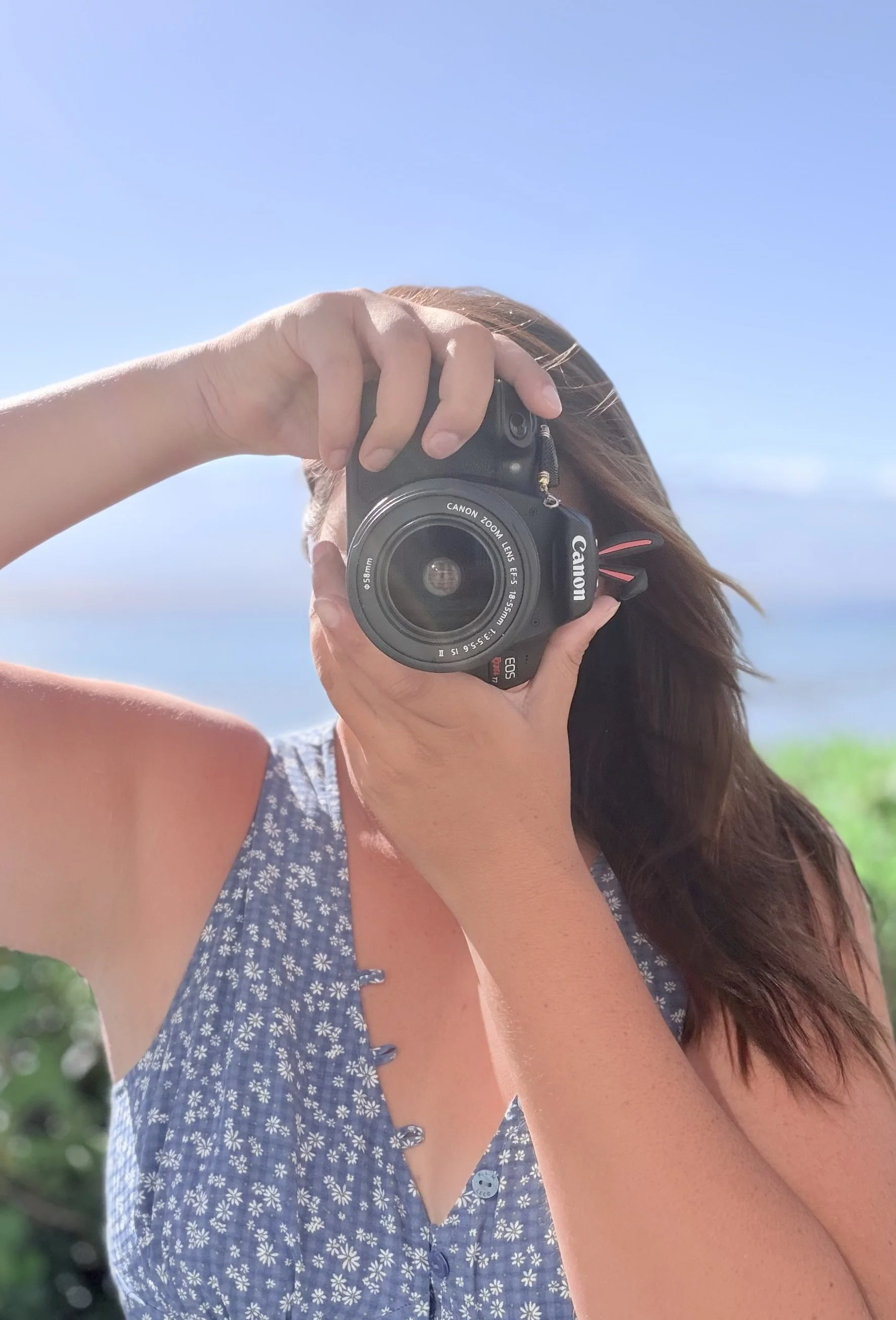 A person holding a Canon camera in front of their face, wearing a blue dress with white floral pattern, outdoors with a clear blue sky.
