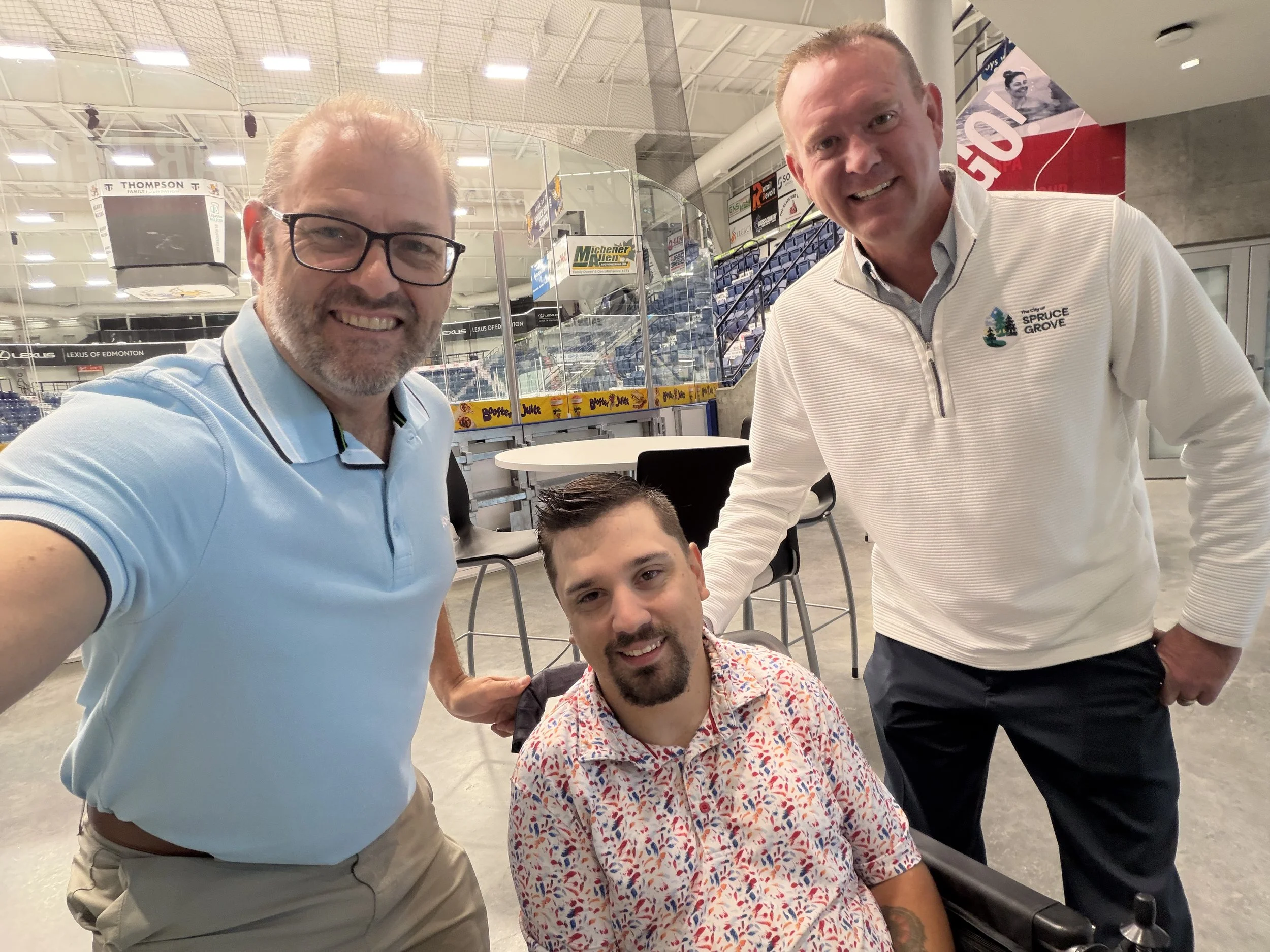 Four men posing together inside a sports arena. One man on the left wearing glasses and a light blue polo shirt. Two men in the middle, one seated and one standing, both smiling. The seated man has a beard and is wearing a colorful patterned shirt. The standing man on the right is wearing a white sweater with 'City of Spruce Grove' logo. The arena has advertisements and seating in the background.