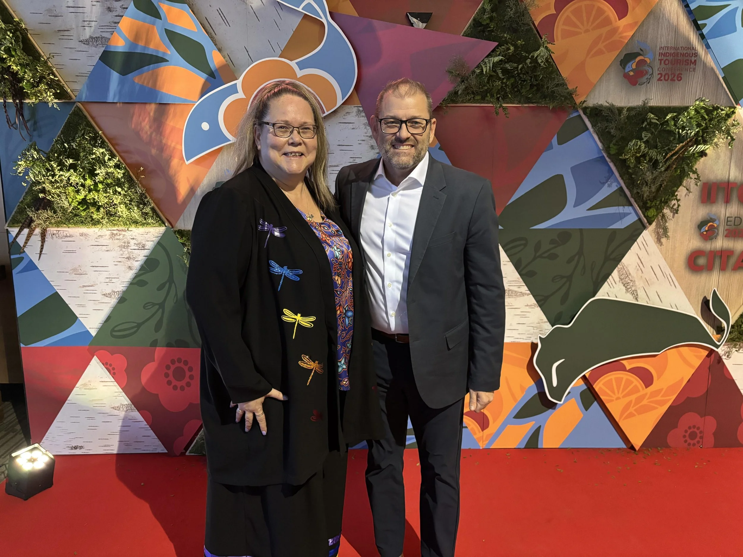 Two people are standing on a red carpet in front of a colorful, geometric backdrop that features signs for the International Indigenous Tourism Conference 2026 and other related themes. They are smiling and dressed in business attire, with the woman wearing glasses and a black jacket, and the man with glasses and a gray suit.