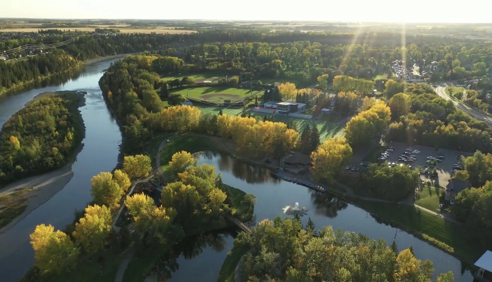 An aerial view of a park with two winding rivers, surrounded by lush green trees with autumn foliage, sports fields, and a parking lot, under a bright sky with sunlight.