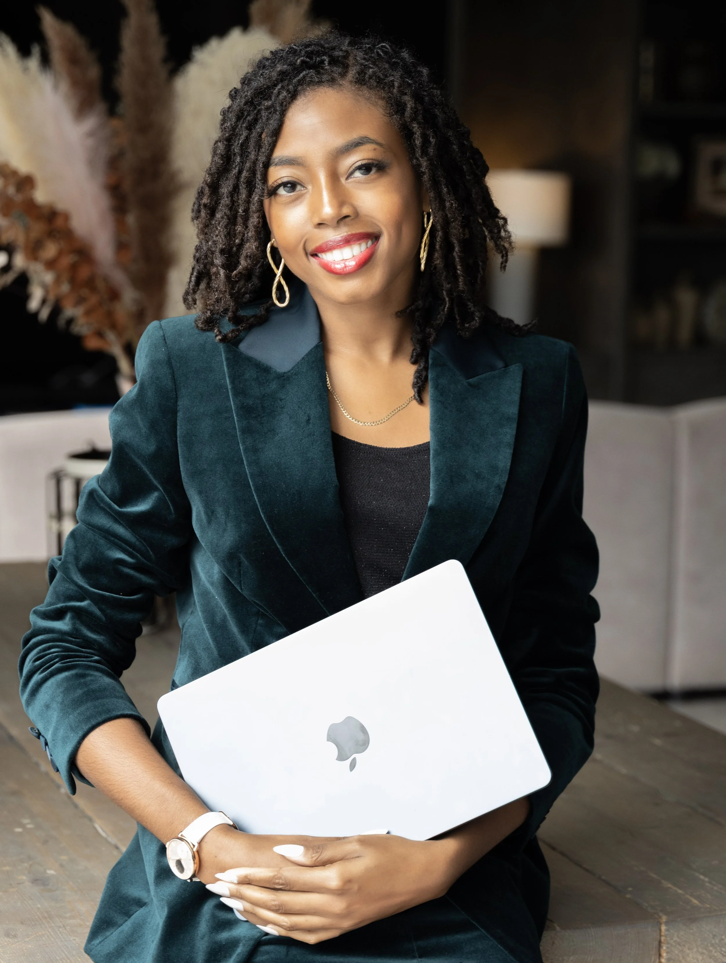 Jenica Leah has dark skin and shoulder-length curly hair is smiling at the camera while holding a silver apple laptop She is wearing a dark teal blazer, gold earrings, and a gold necklace.