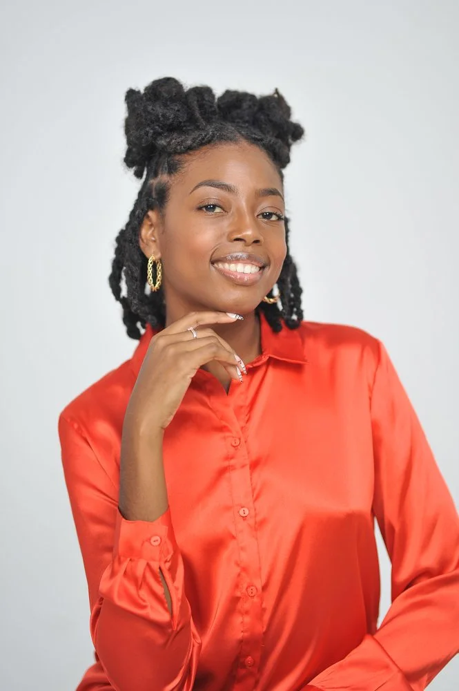 Jenica Leah, a woman with dark skin and styled dreadlocks smiling and posing with her hand near her chin, wearing a red satin blouse and gold hoop earrings.