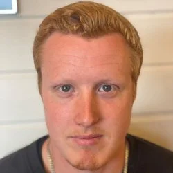 Nærbilde av en mann med lys hud, rødt hår og blå øyne, iført svart t-skjorte, med en hvit trevegg i bakgrunnen.