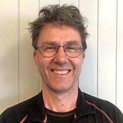 Et bilde av en mann med brunt, krøllete hår og briller, smilende, mot en lys bakgrunn.