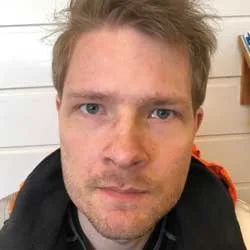 Ung mann med lyst hår og blå øyne, iført svart jakke, foran en hvit vegg.