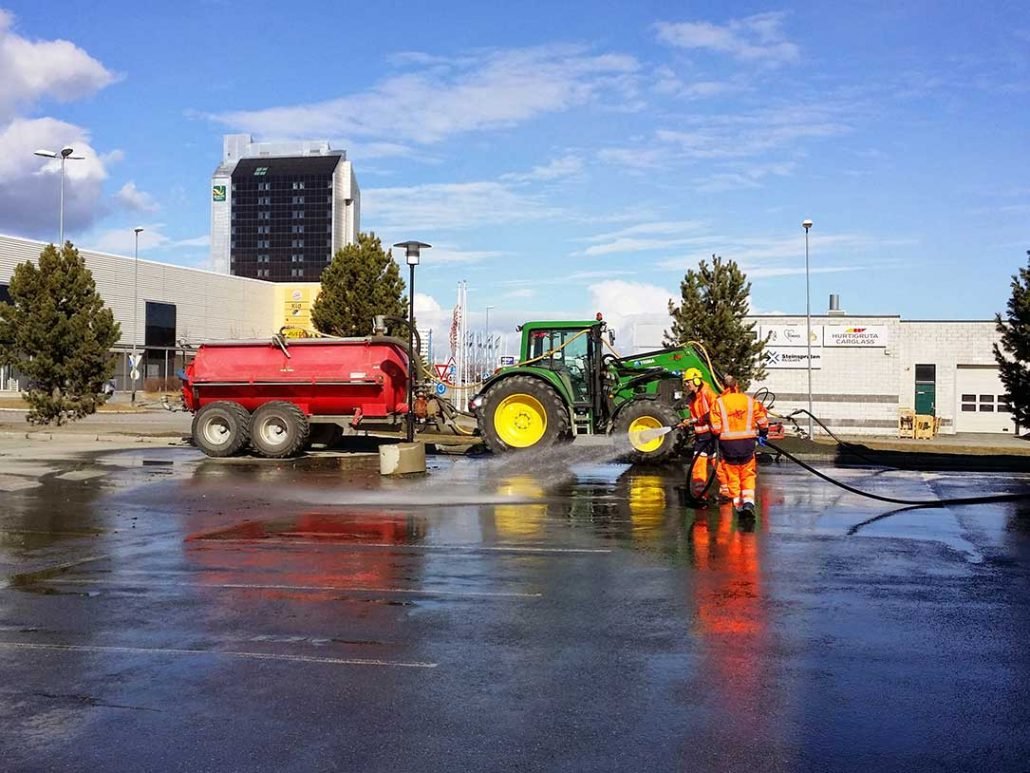 To veiledere i orange arbeidstøy rengjør asfalten med høytrykksspyler mens en traktor trekker en vannvogn på tom parkeringsplass, med universitetsbygg i bakgrunnen og en byhimmel.