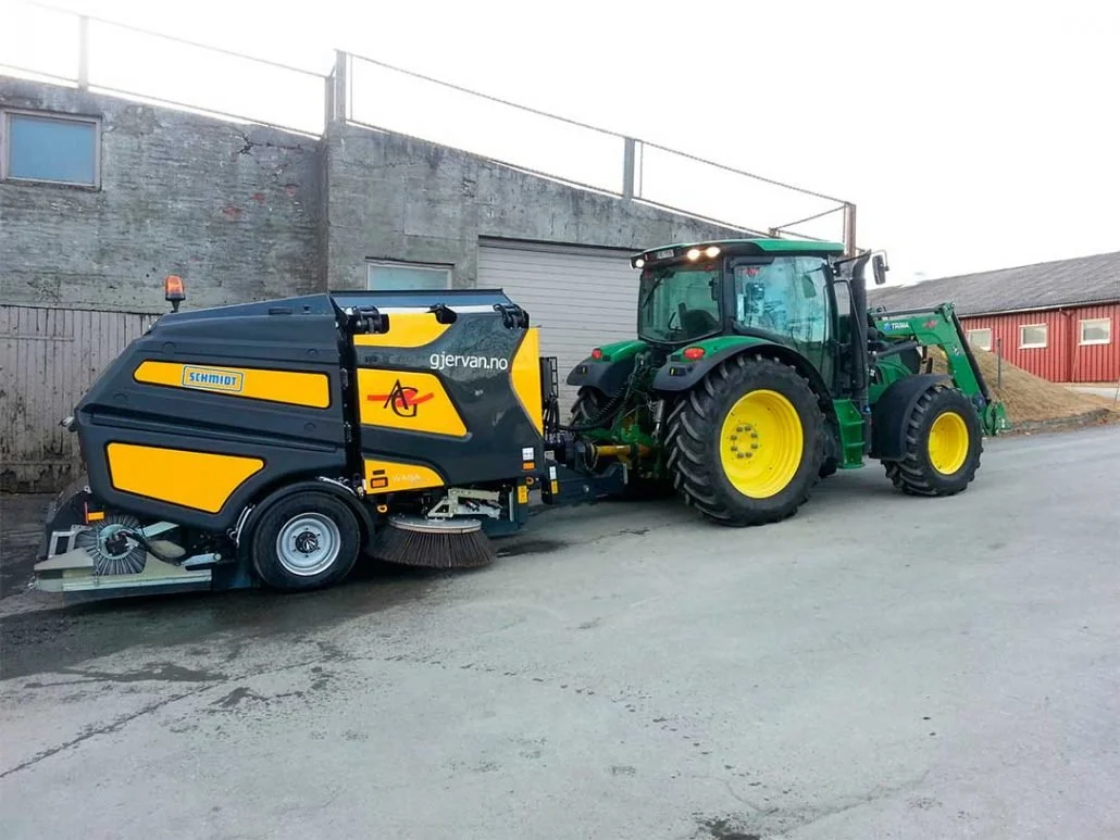 En grønn traktor med gule hjul som er koblet til en svart og gul gatefeier maskin, parkert ved en bygning.