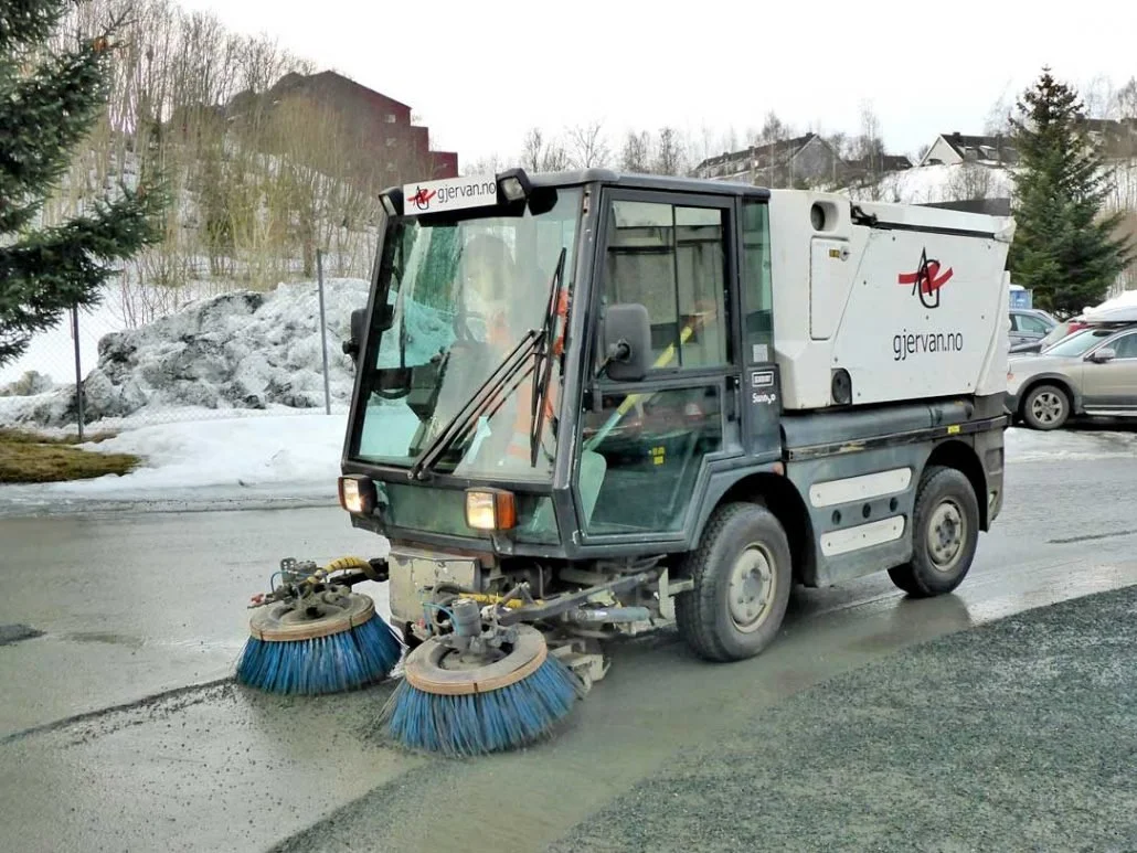 Liten gatefeie med blå børster som renser asfalten, parkert på en vei med snø og kalde omgivelser.