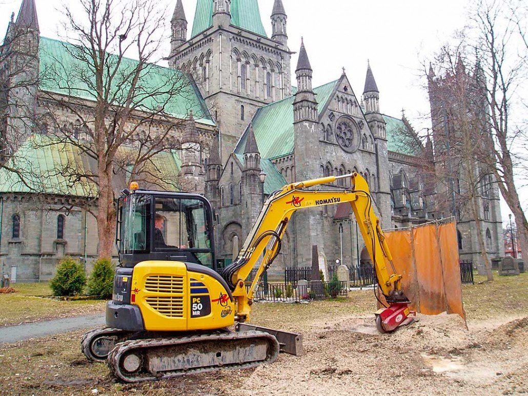En liten gravemaskin foran en stor katedral med grønne tak, omgitt av trær og en liten sikkerhetsgardin.