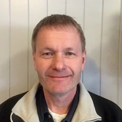 Portrett av en mann med lyst hår og smil, iført en svart og hvit jakke, stående foran en lysegrå vegg.