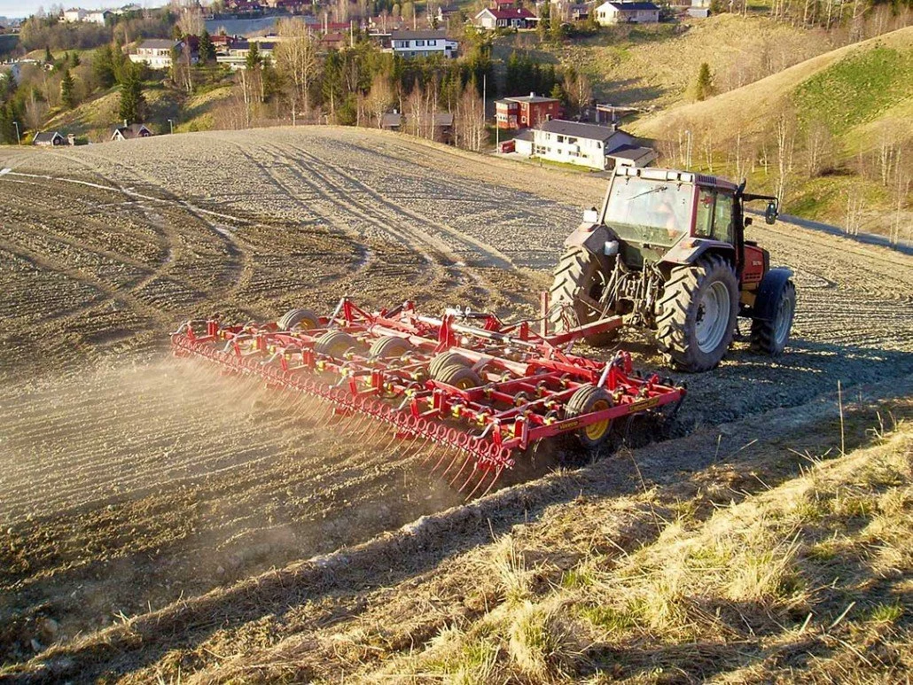 En rød traktor med jordbehandlingsutstyr som pløyer jord i en åker, med boligområder i bakgrunnen.