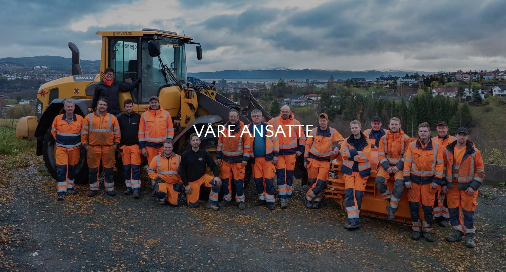 En gruppe arbeidere i orange og blå arbeidsklær står foran en stor gravemaskin med utsikt over en by og fjell i bakgrunnen, med teksten 'VÅRE ANSATTE' over bildet.