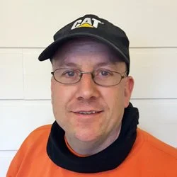 En mann iført sort caps og oransje arbeidsoverall poserer foran en hvit vegg.