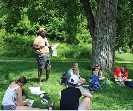 A group of people sitting and standing under a large tree in a park, reading and relaxing on grass.