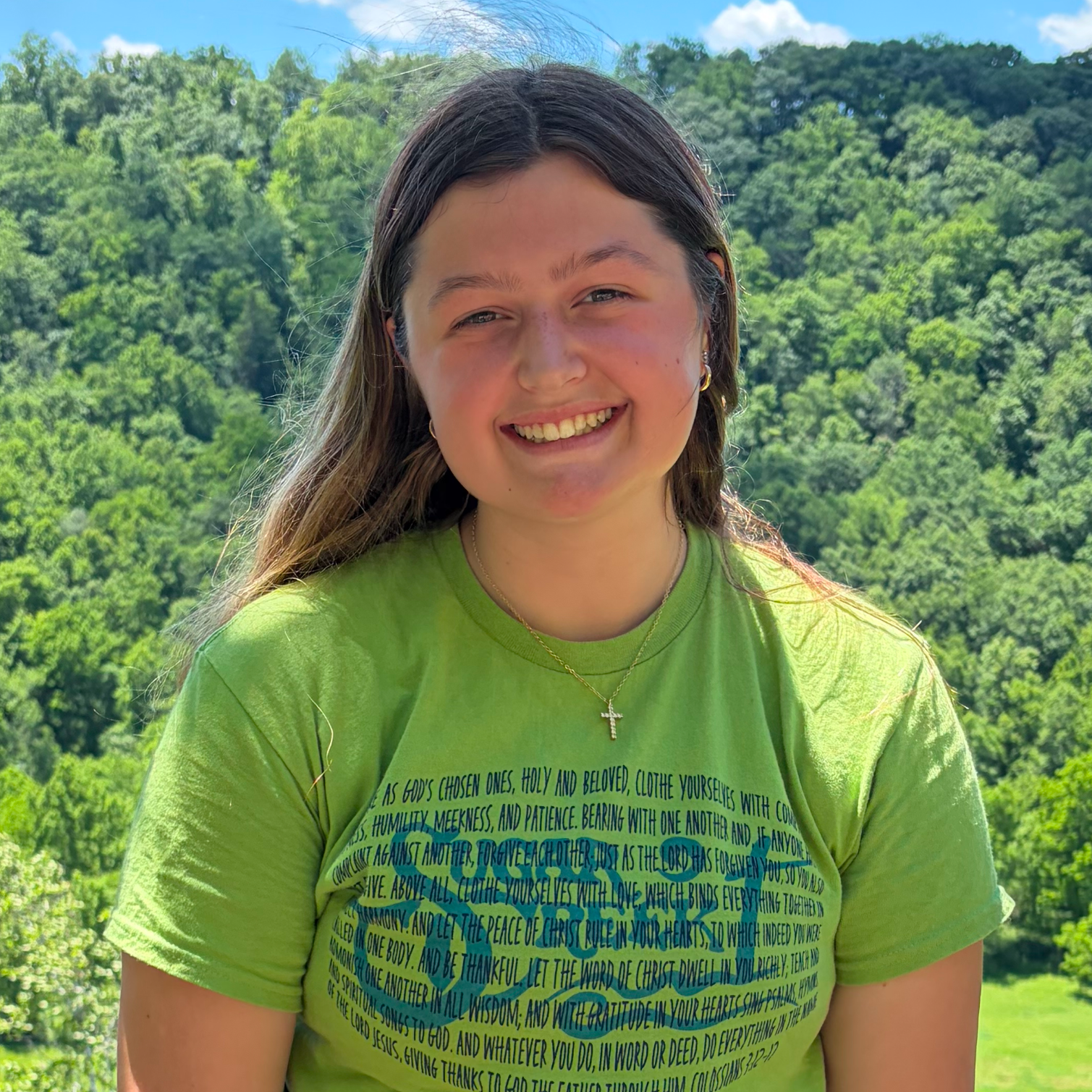 A young woman smiling outdoors with green trees and a blue sky in the background. She is wearing a bright green T-shirt with text on it and a silver cross necklace.
