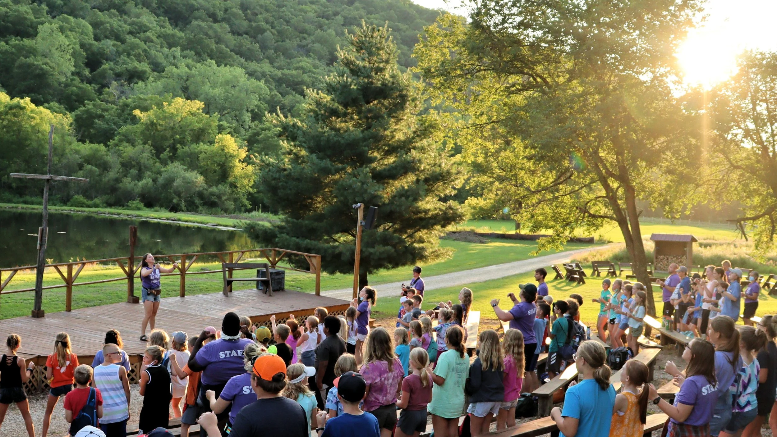 A large outdoor gathering of people, including children and adults, watching a performer on a wooden stage near a pond with green trees and hills in the background, during the late afternoon or early evening sunlight.