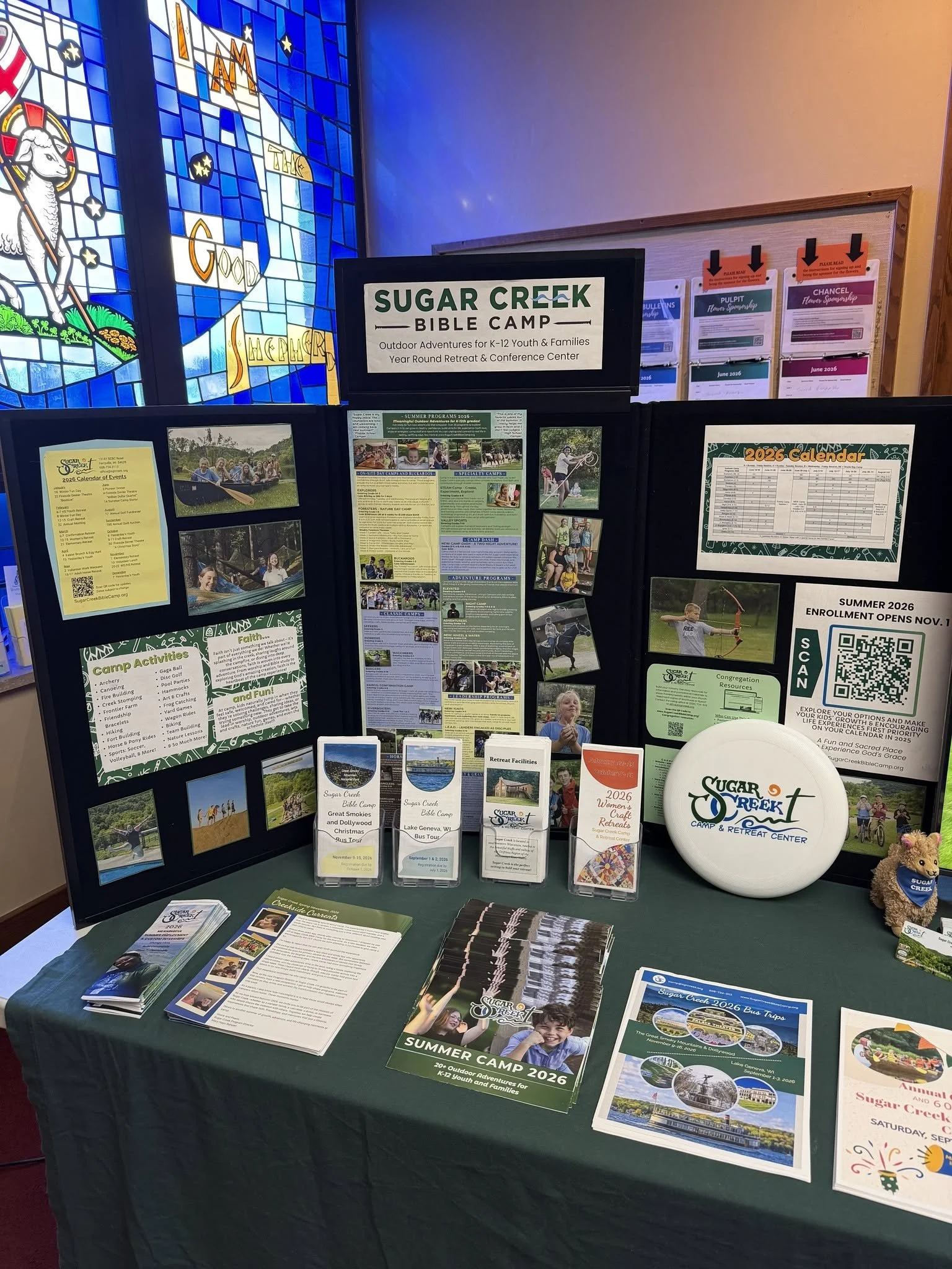 Information booth for Sugar Creek Bible Camp with flyers, booklets, and brochures about summer 2026 outdoor adventures for youth and families, and a stained glass window of a lamb and a rainbow in the background.