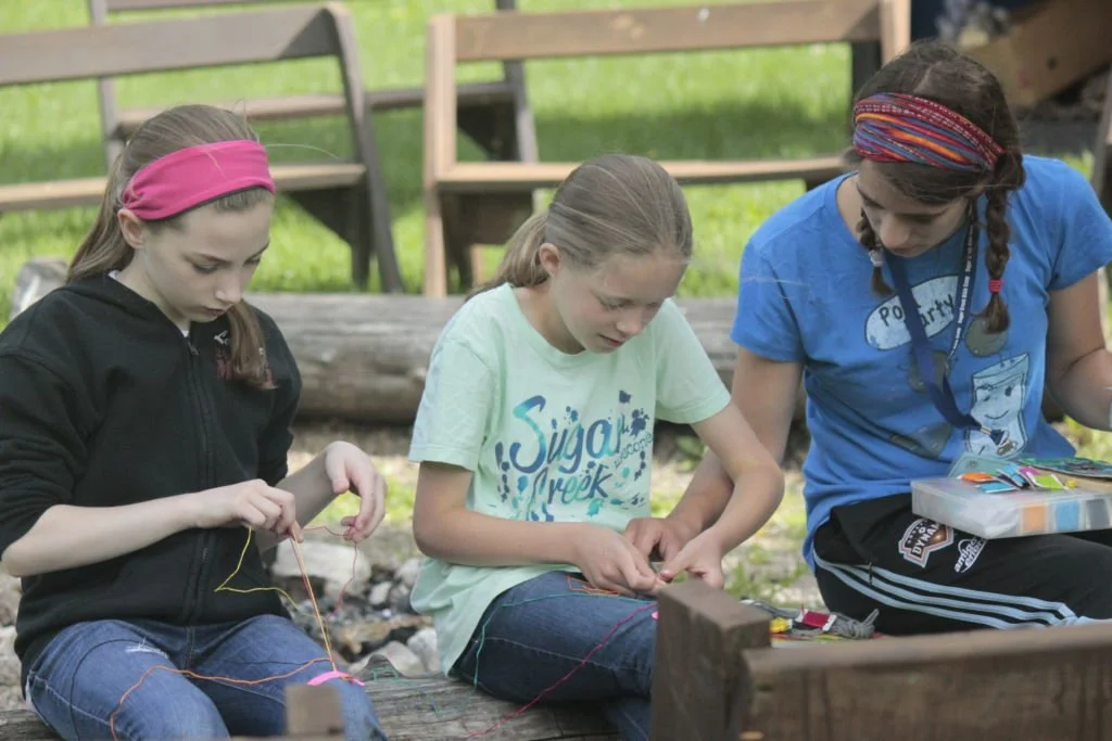 Three girls sitting outdoors on a log, working on a craft project with string, in a wooded area with chairs in the background.