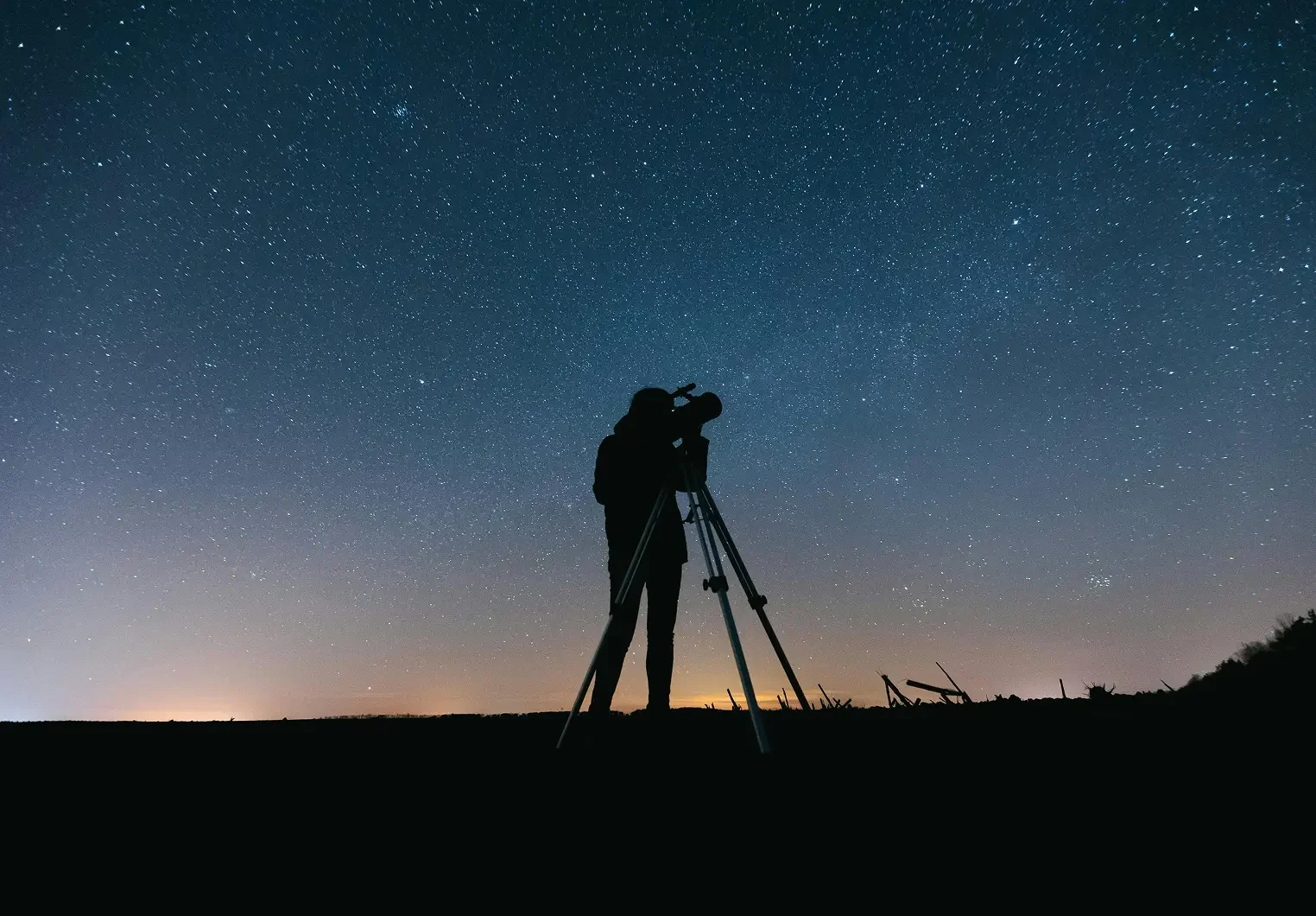 Silhouette of a person using a telescope at night under a starry sky with a faint glow on the horizon.