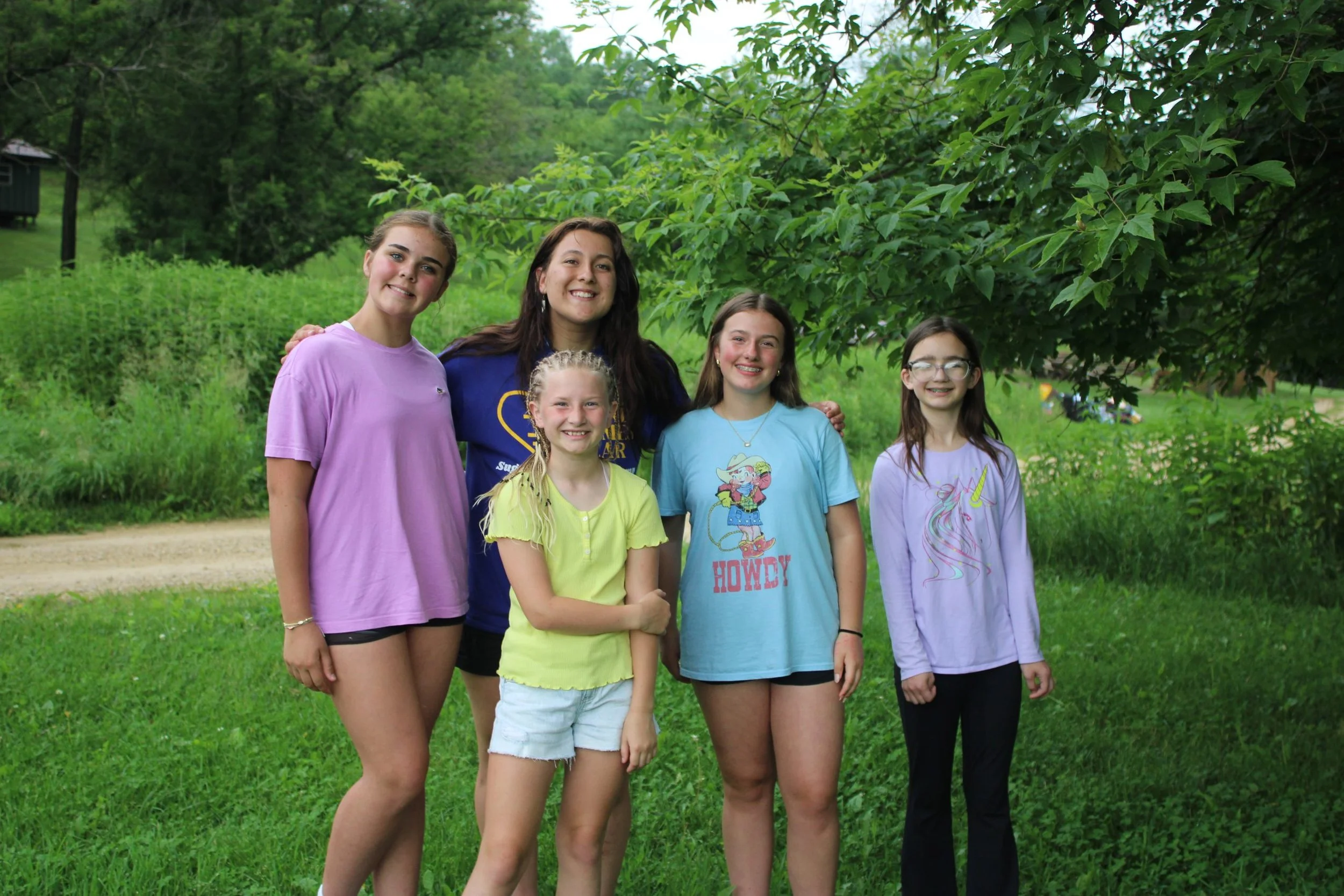 Six girls standing outdoors on green grass, smiling, with trees and shrubs in the background.