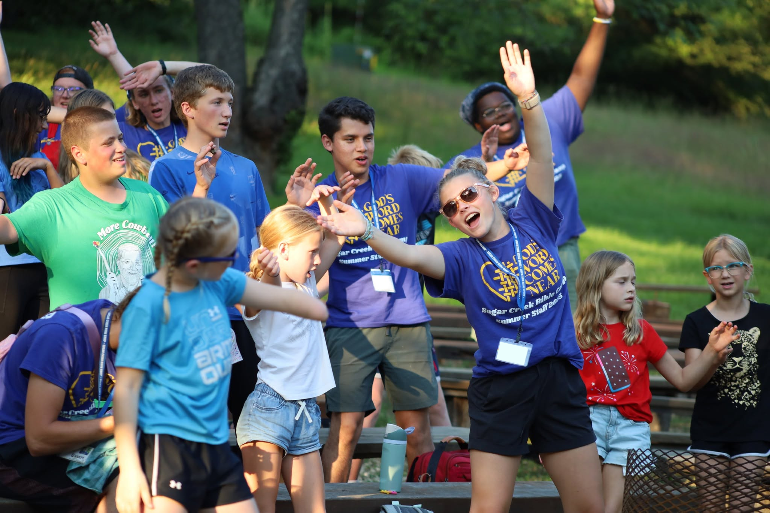 A group of children and teenagers outdoors, some raising their hands and singing or talking, participating in an organized event or camp in a park-like setting with trees and green grass.