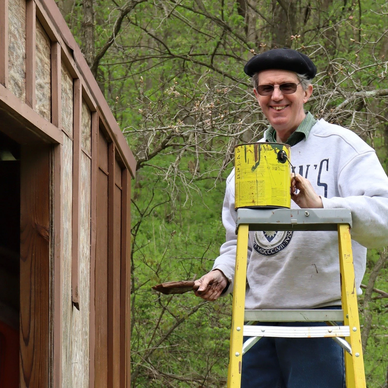A man on a yellow ladder painting the exterior of a wooden shed with a paintbrush. He is wearing sunglasses, a black beret, a white sweatshirt, and a green shirt underneath. The background features trees with budding leaves.