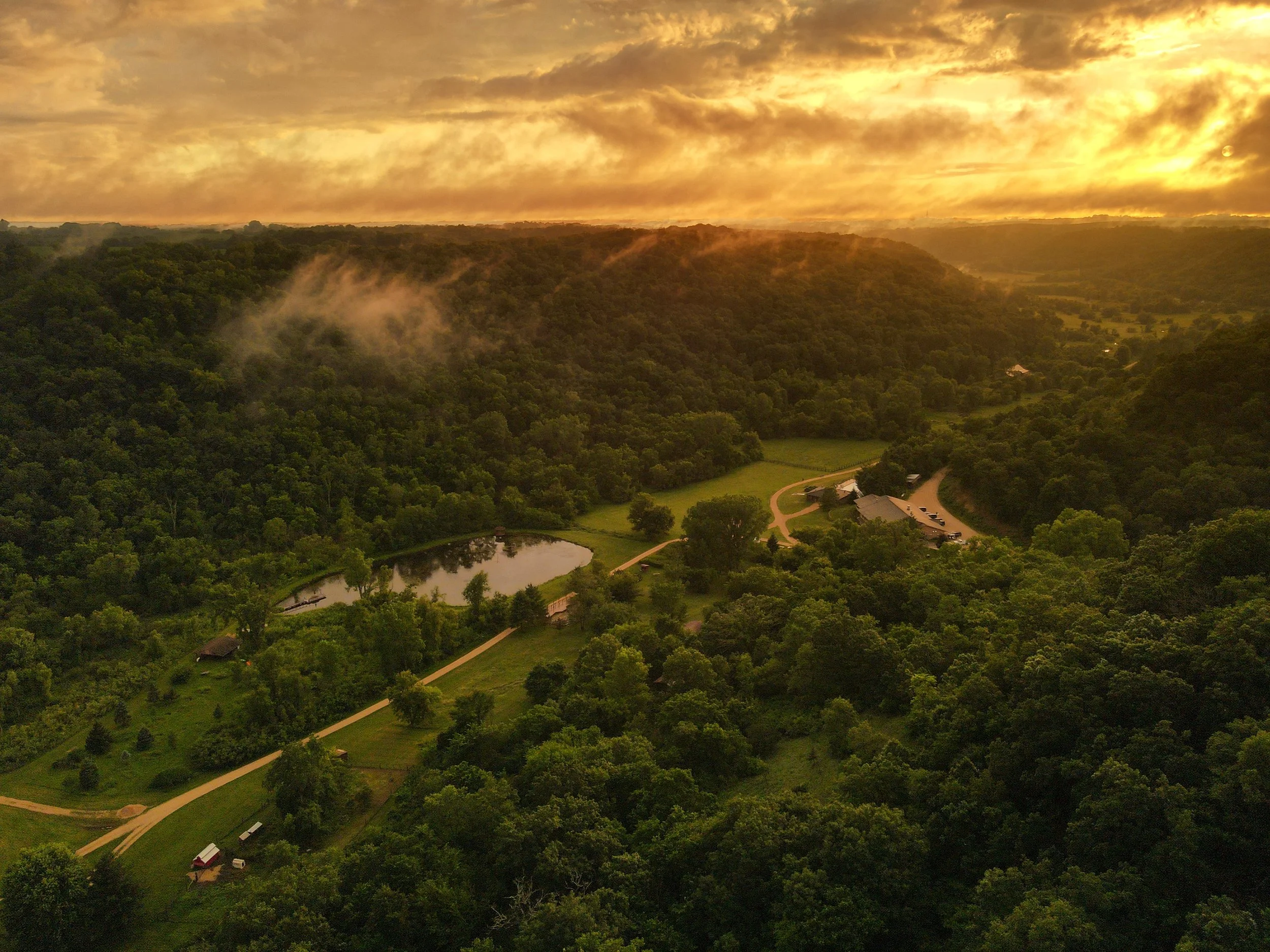 Overhead view of a lush green landscape with trees, a small pond, scattered buildings, roads, and a vibrant sunset with orange and yellow clouds.