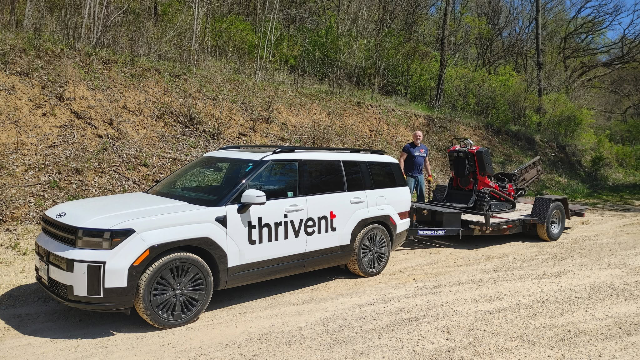 A white SUV with the word 'thrivent' on the side is towing a trailer carrying a small tracked excavator, with a man standing nearby on a dirt road with a wooded hillside in the background.