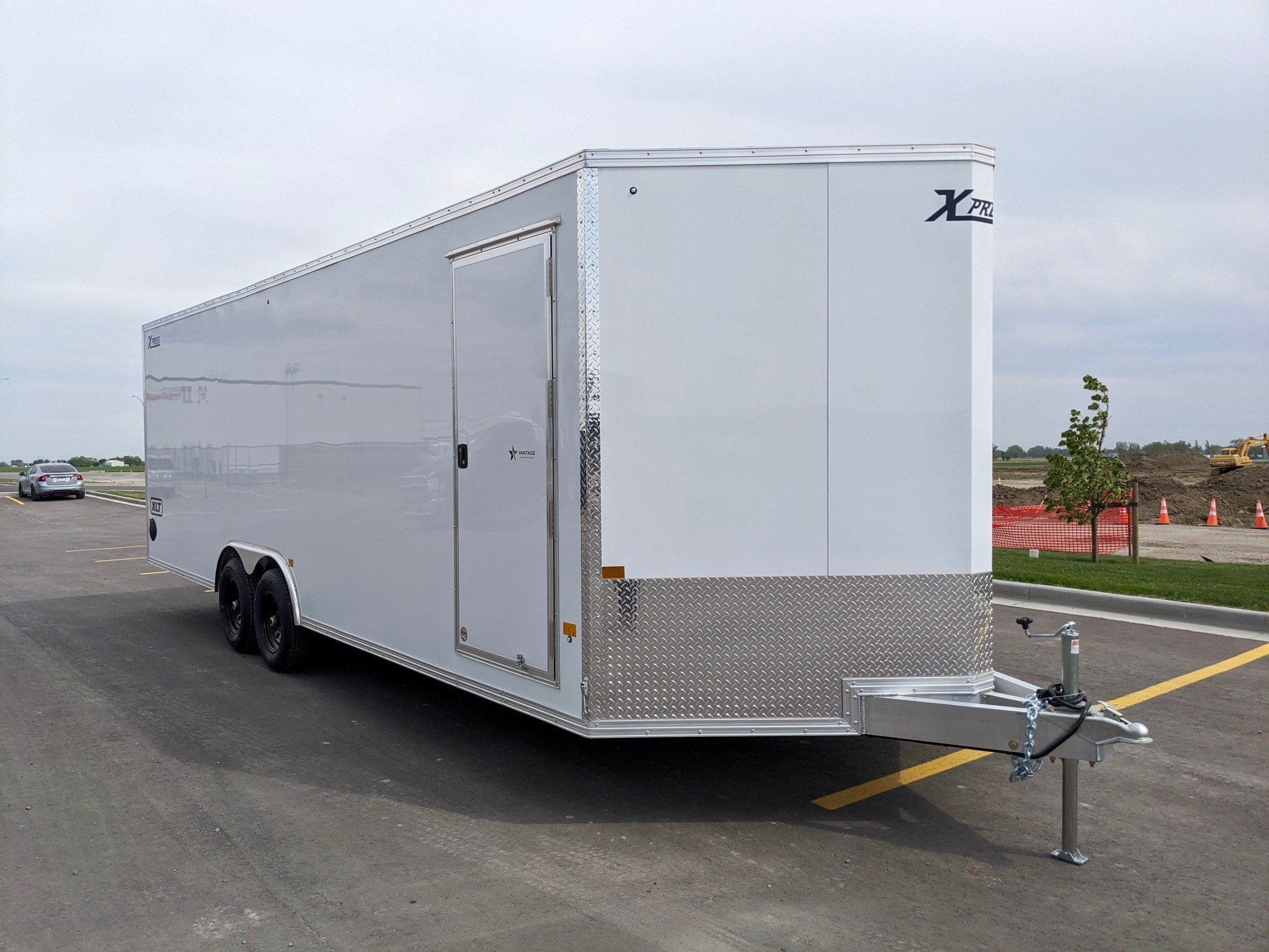 A white enclosed trailer with a side door and a logo in the upper right corner, parked on asphalt in a lot with a car and construction area nearby.