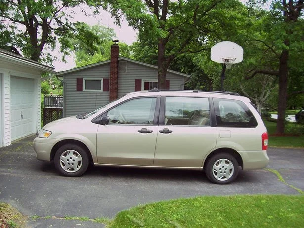 A beige minivan parked on a driveway in front of a house with a basketball hoop and green trees.