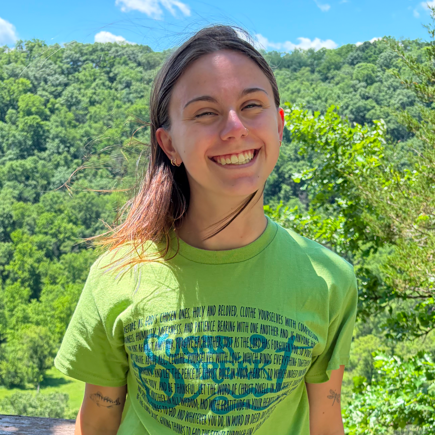 A smiling young woman with dark hair, light skin, and a nose piercing, wearing a green t-shirt with blue text and standing outdoors against a lush green landscape and a blue sky with clouds.