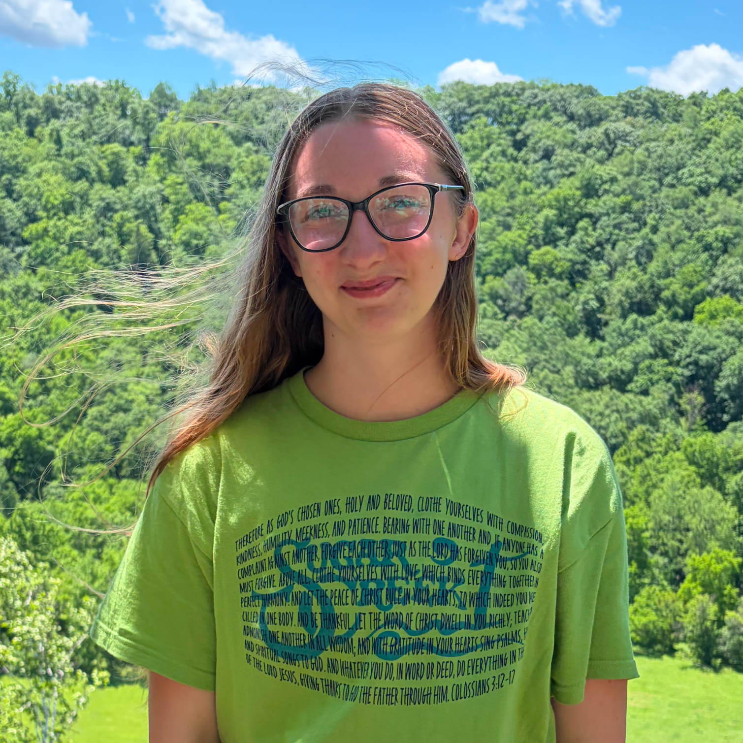 Young woman with long hair and glasses standing outdoors with lush green trees and a blue sky in the background, wearing a bright green T-shirt with text and a circular design.