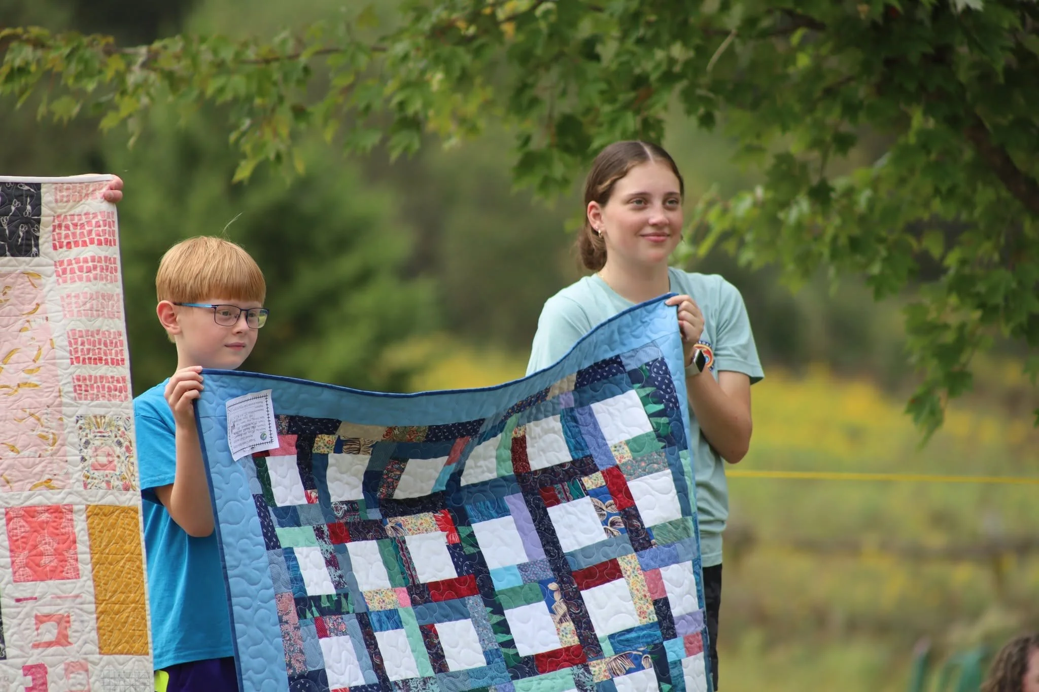 Two children and an adult holding a colorful quilt in an outdoor setting with green trees in the background.