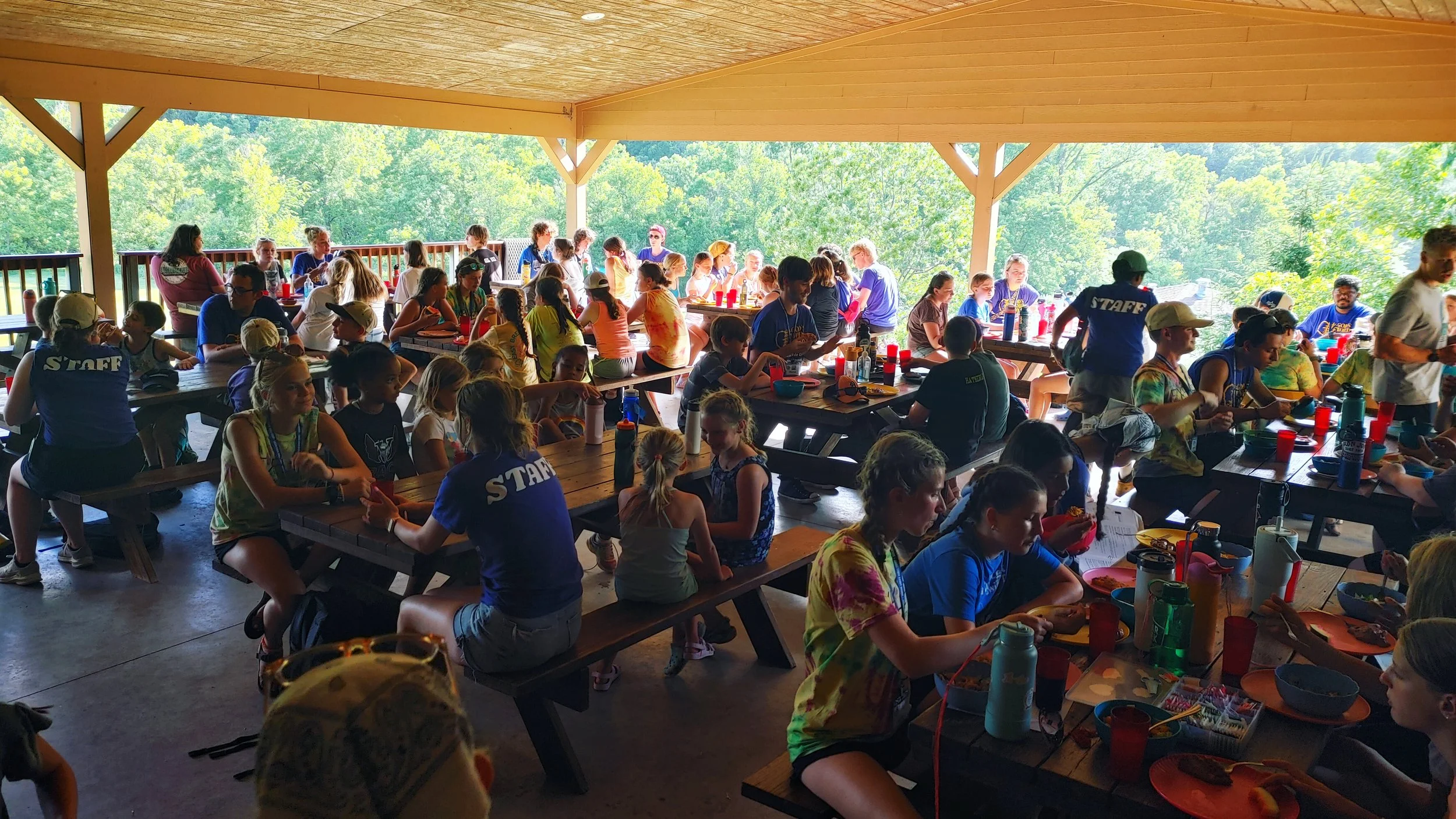 A large group of children and teenagers sitting at wooden picnic tables underneath a covered outdoor pavilion, surrounded by green trees, with some adults supervising.