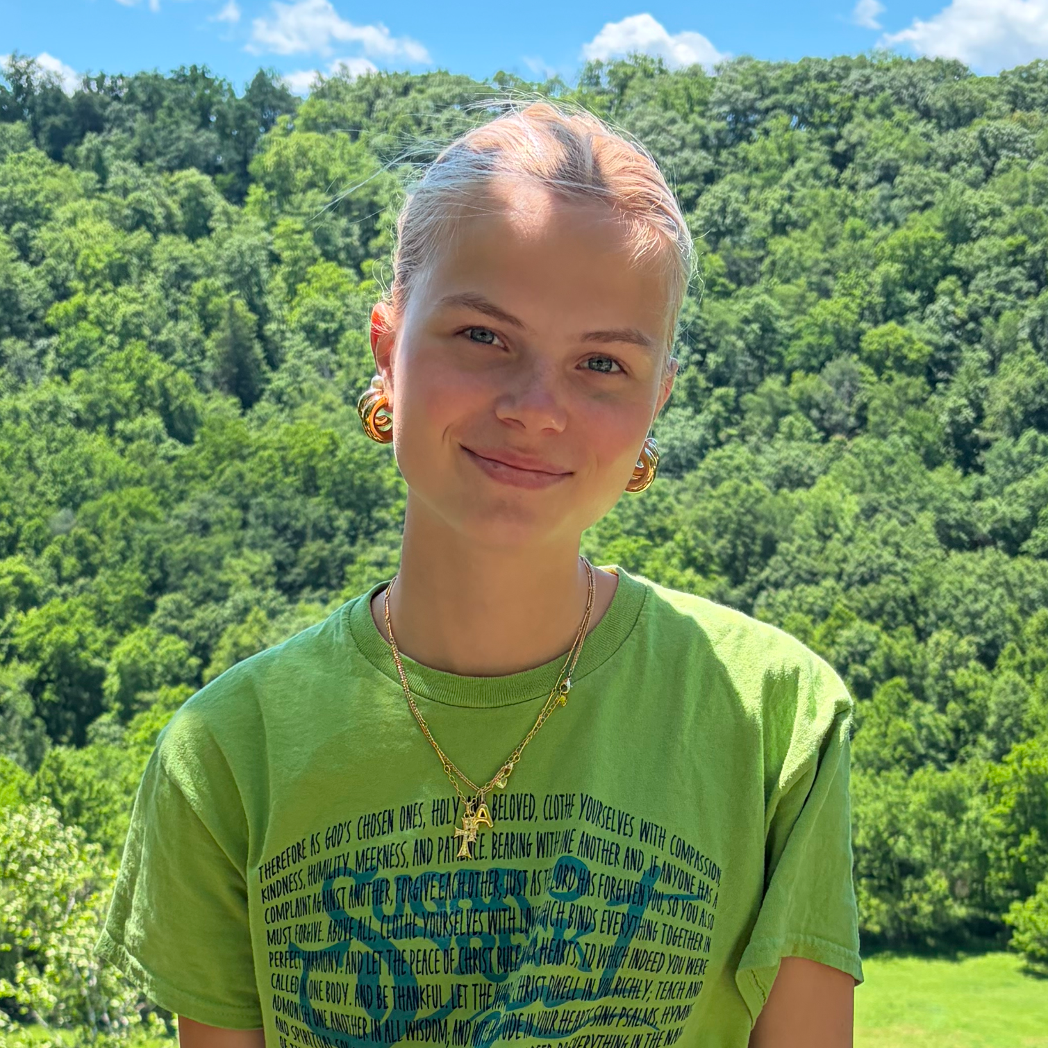 A young woman with blonde hair tied back, wearing gold hoop earrings and layered necklaces, smiling outdoors with a lush green forest in the background under a blue sky with clouds.