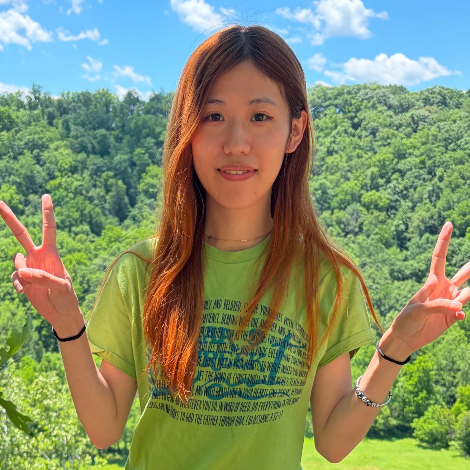 A young woman with long reddish hair smiling and making peace signs with both hands outdoors in front of trees and a blue sky with clouds, wearing a lime green t-shirt with colorful text.