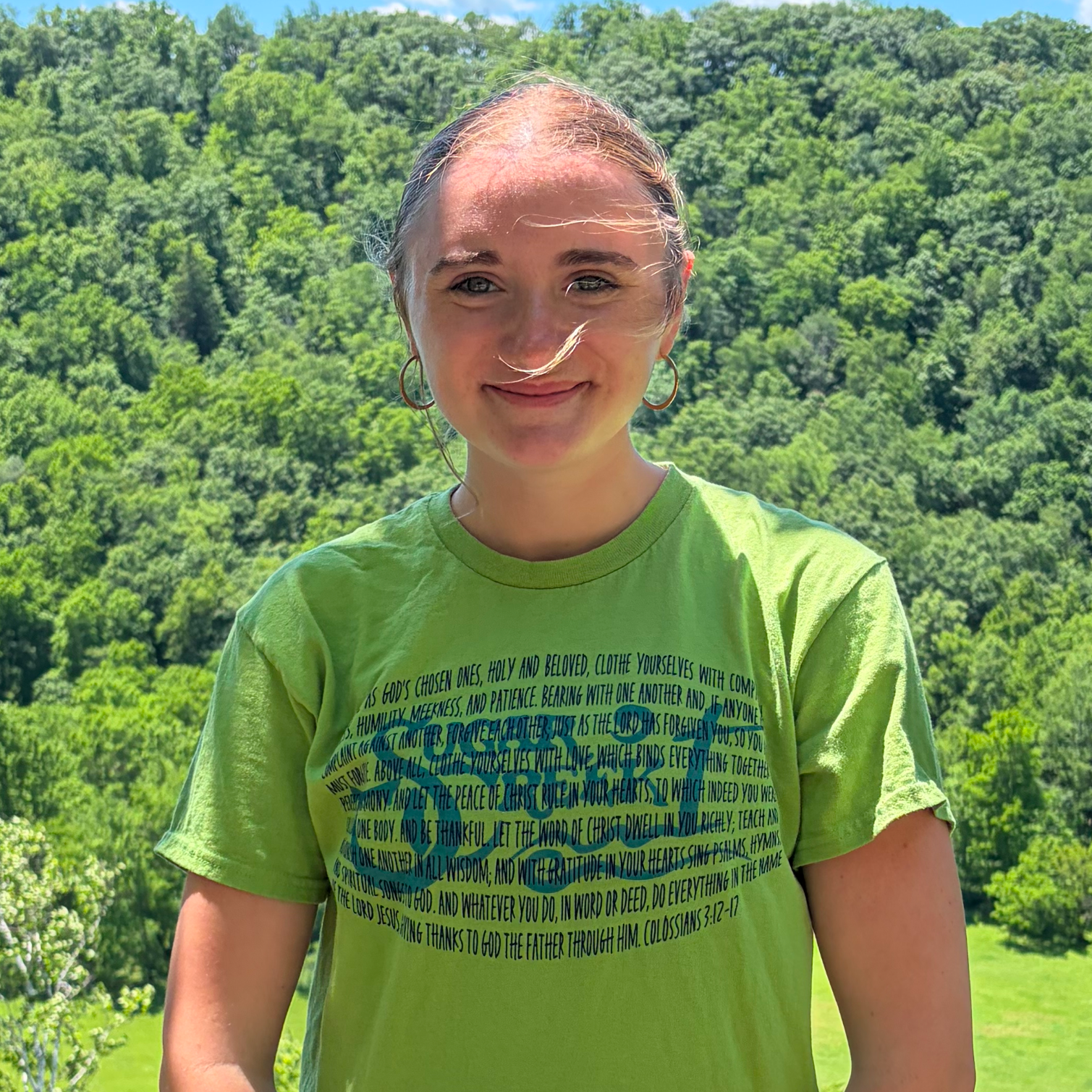 A young woman standing outdoors, wearing a bright green T-shirt with text on it, and earrings, with a background of lush green trees and hills under a clear sky.