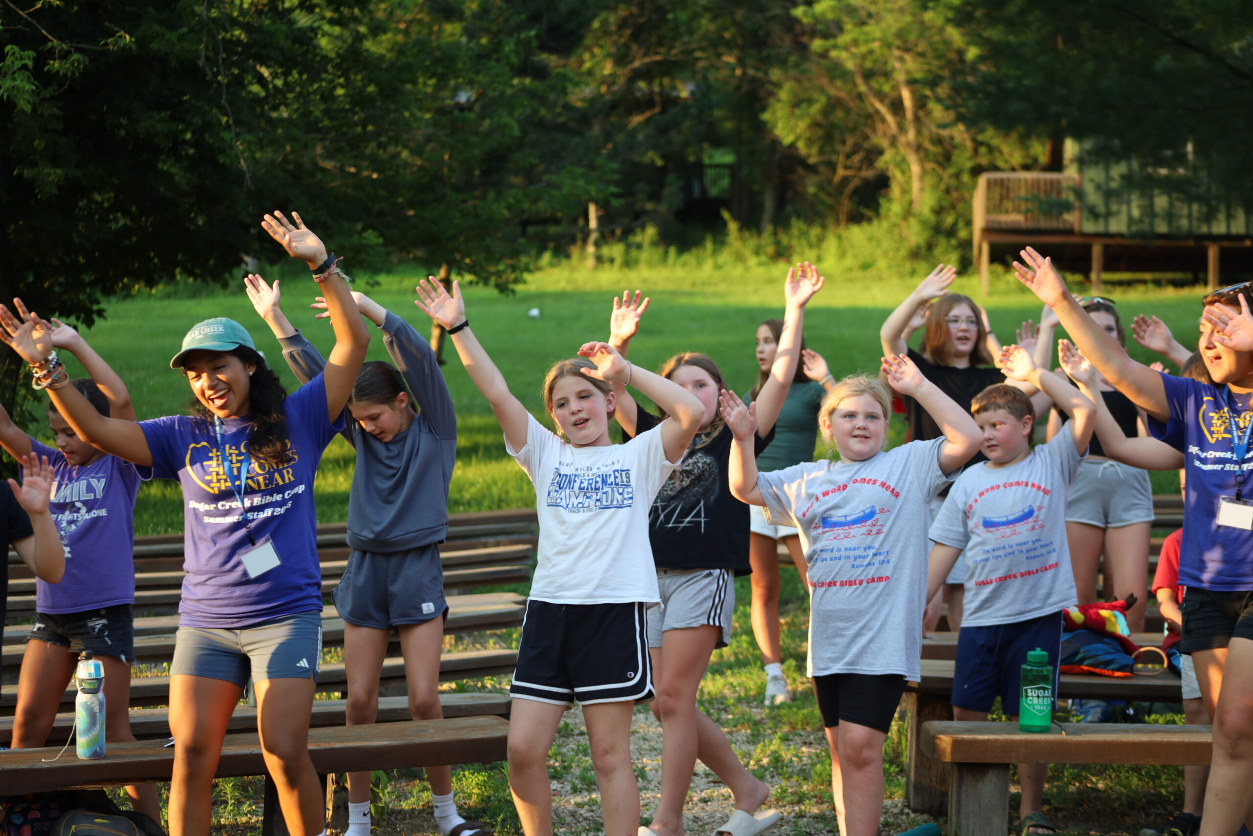 A group of children participating in an outdoor activity or camp, raising their hands while standing on wooden steps in a park-like setting with green trees and grass.