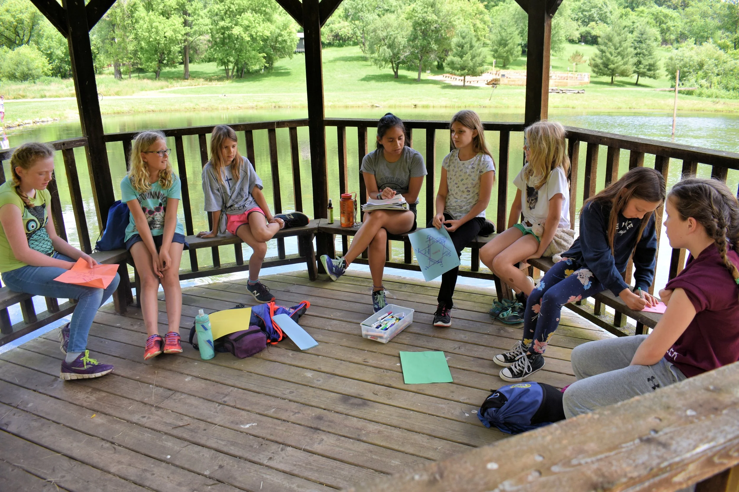 A group of girls sitting on the wooden floor of a gazebo near a lake, engaging in arts and crafts activities with papers, markers, and supplies, surrounded by lush green trees and a peaceful outdoor setting.