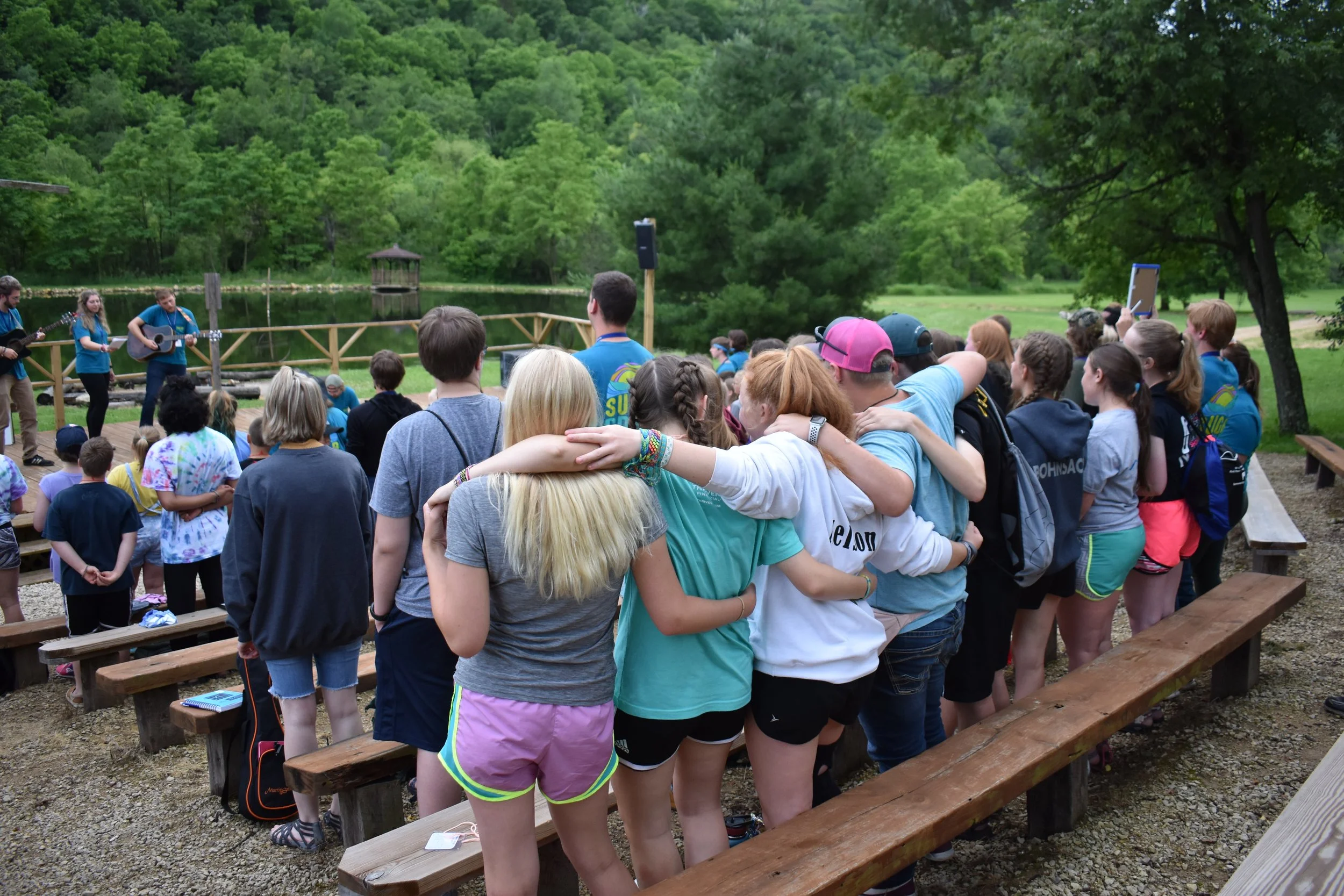 A large group of children and teenagers gathered outdoors around a stage, listening to a band performing with guitars near a pond and surrounded by trees.