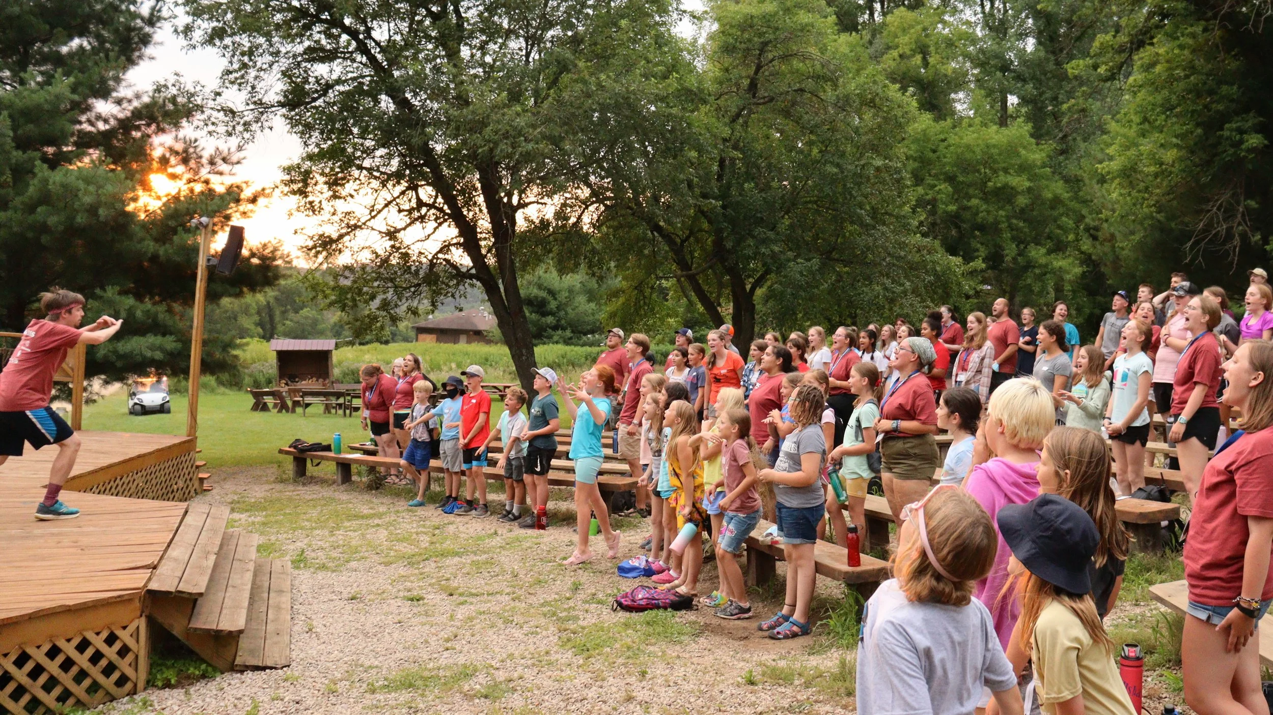 A large group of children and adults gathered outdoors on wooden benches, watching a performer on a small stage at sunset surrounded by trees.