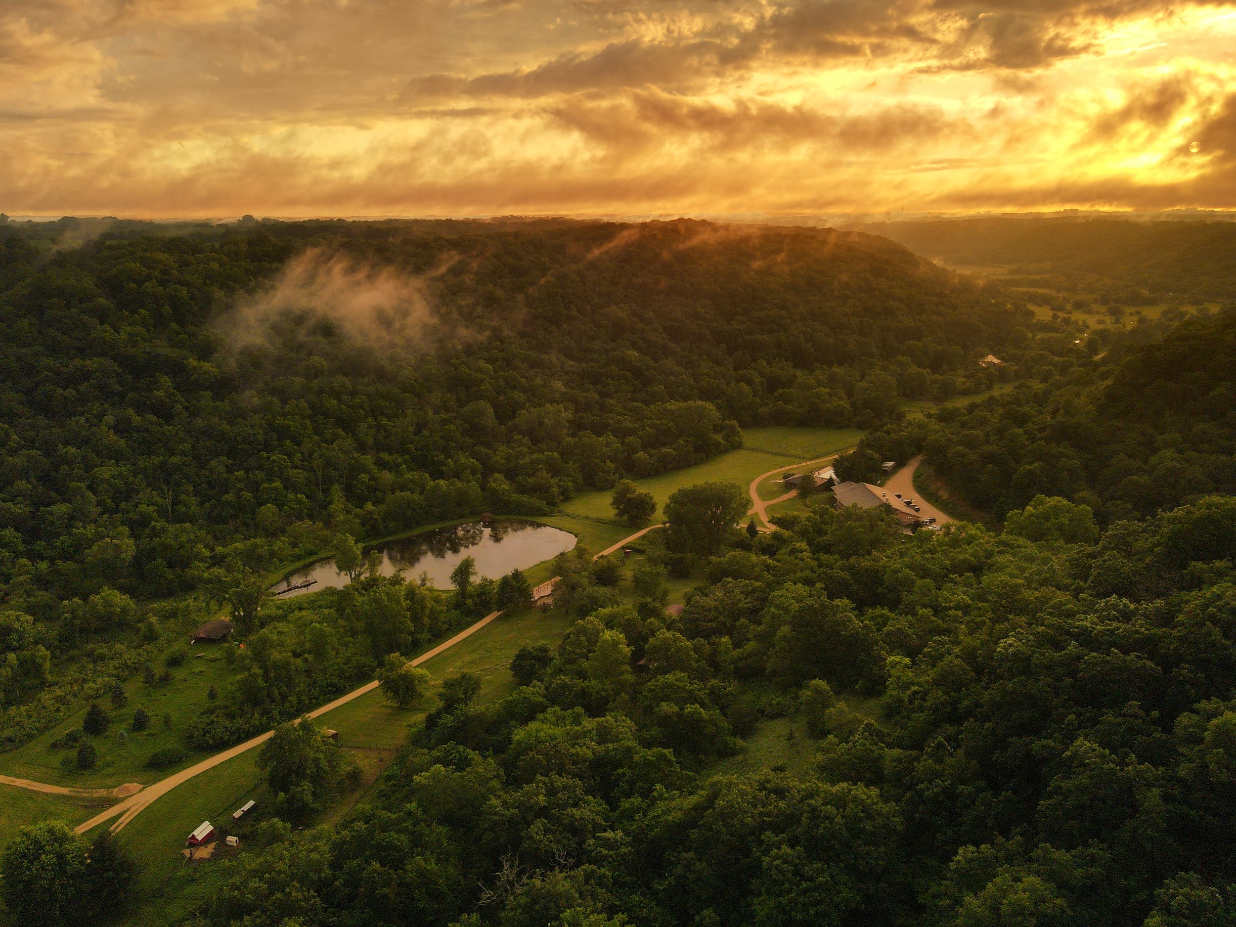 A scenic view of a forested valley at sunset with a dramatic yellow and gray cloud-filled sky, a small pond, winding paths, and a few cabins visible in the landscape.