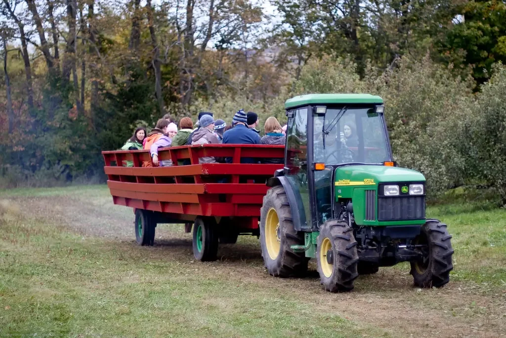 A green John Deere tractor pulling a wooden red wagon filled with children through a grassy field with trees in the background.