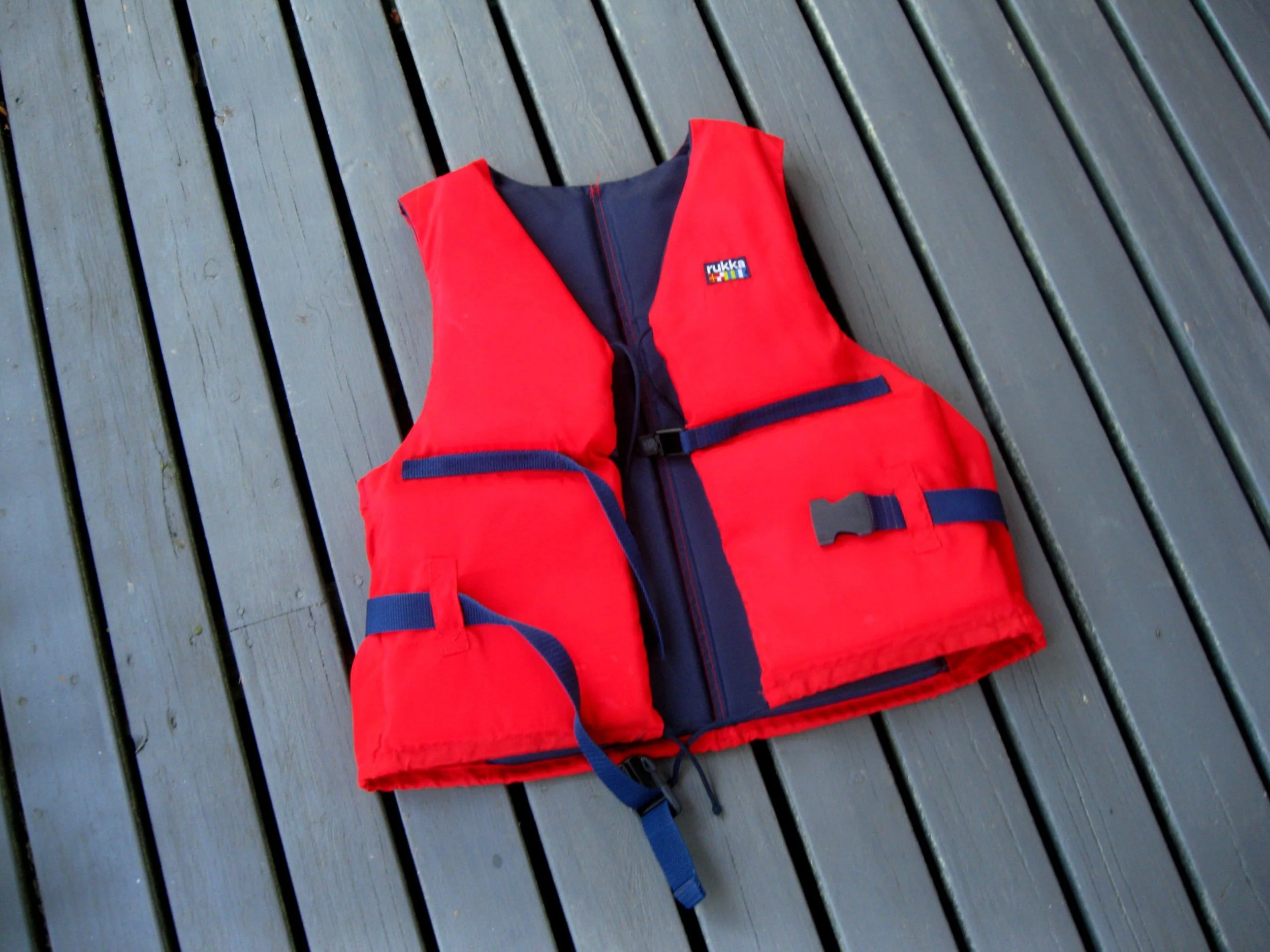 Red life jacket with black straps and a small multicolored label lying on a gray wooden deck.